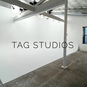Interior of a photography studio with white walls, concrete floor, and large window, featuring black text 'TAG STUDIOS' on the wall.
