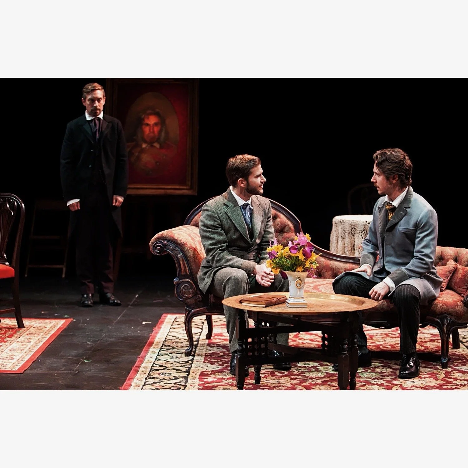 Two men in vintage suits sitting on a floral sofa, engaged in conversation, with a vase of flowers on a table between them, while another man in a dark suit stands behind them, and a portrait hangs on the wall in the background.