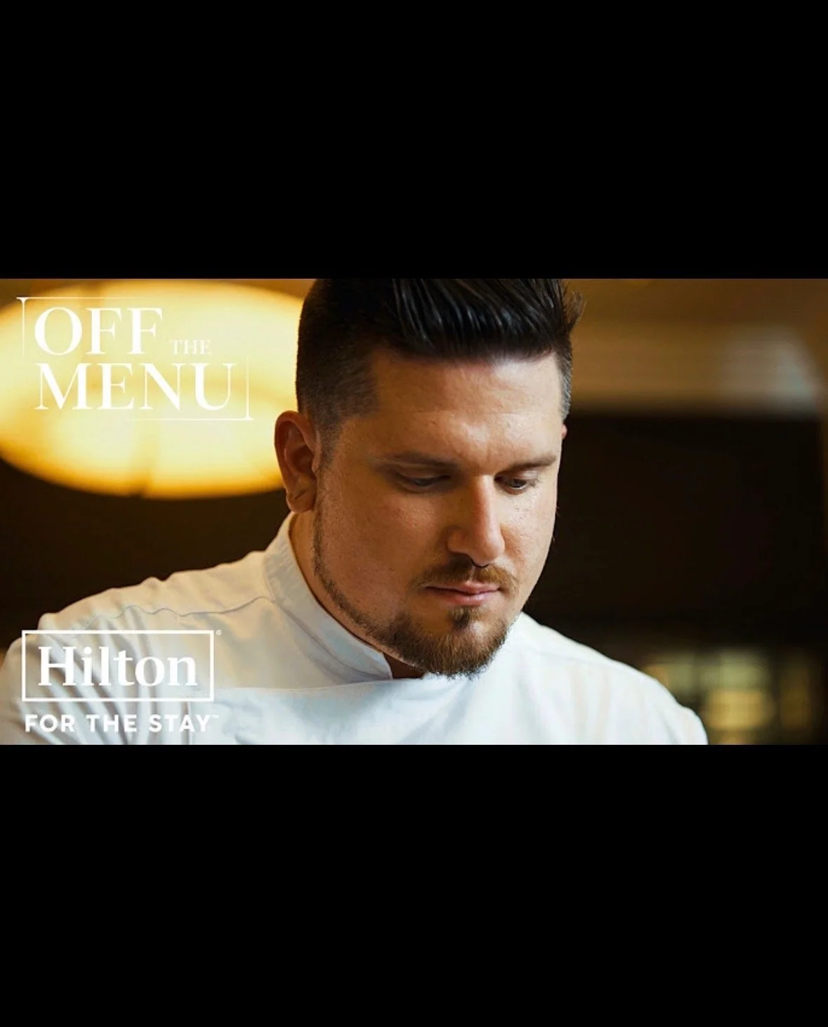 Young male chef with dark hair and beard, looking down, in a dining area with warm lighting.