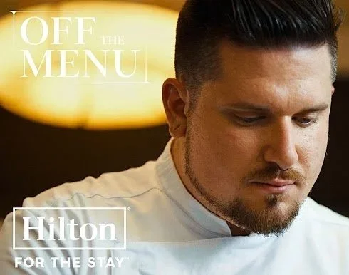 Close-up of a male chef in a white uniform looking down, with the Hilton logo and text 'OFF THE MENU' and 'FOR THE STAY' in the background.
