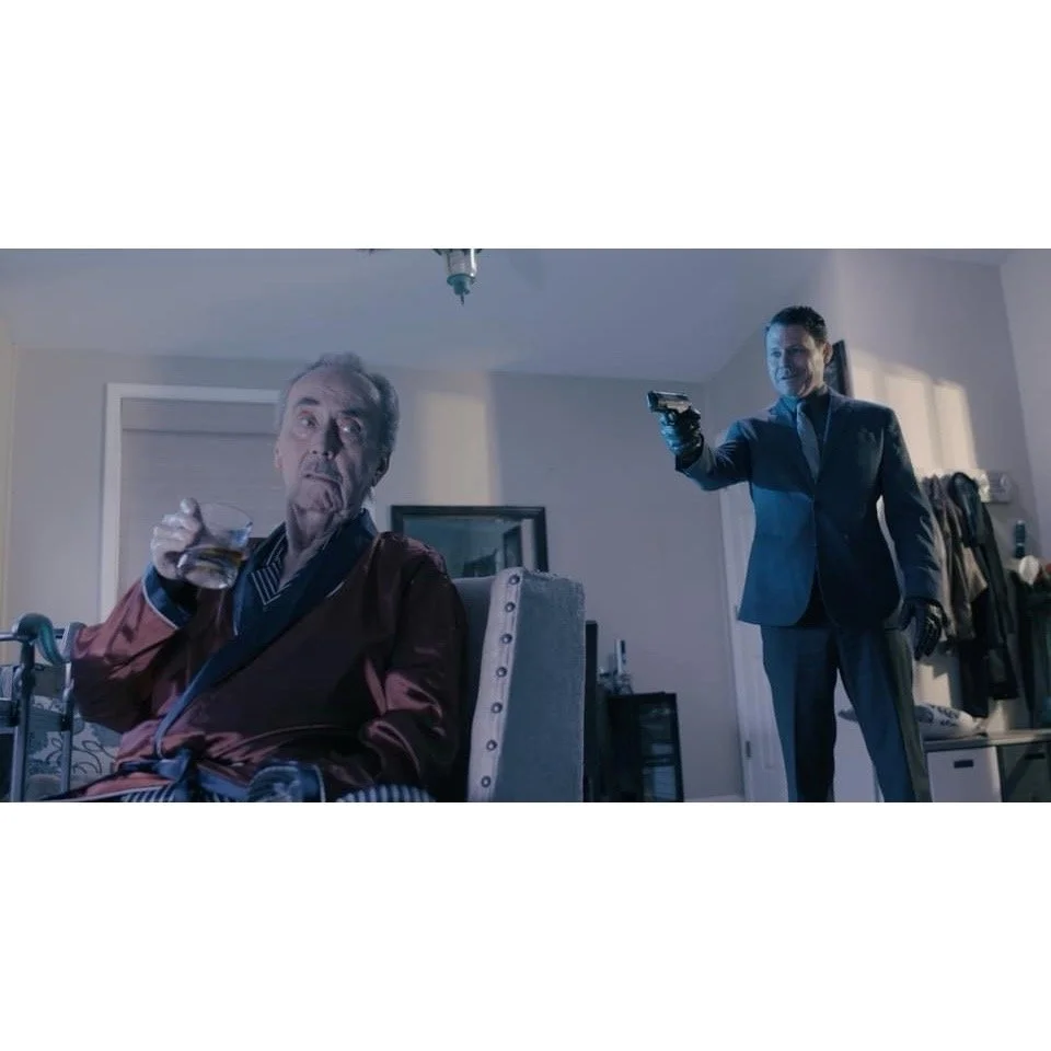 A man in a suit points a gun at another older man sitting in a chair, who is holding a drink and appears surprised or alarmed.