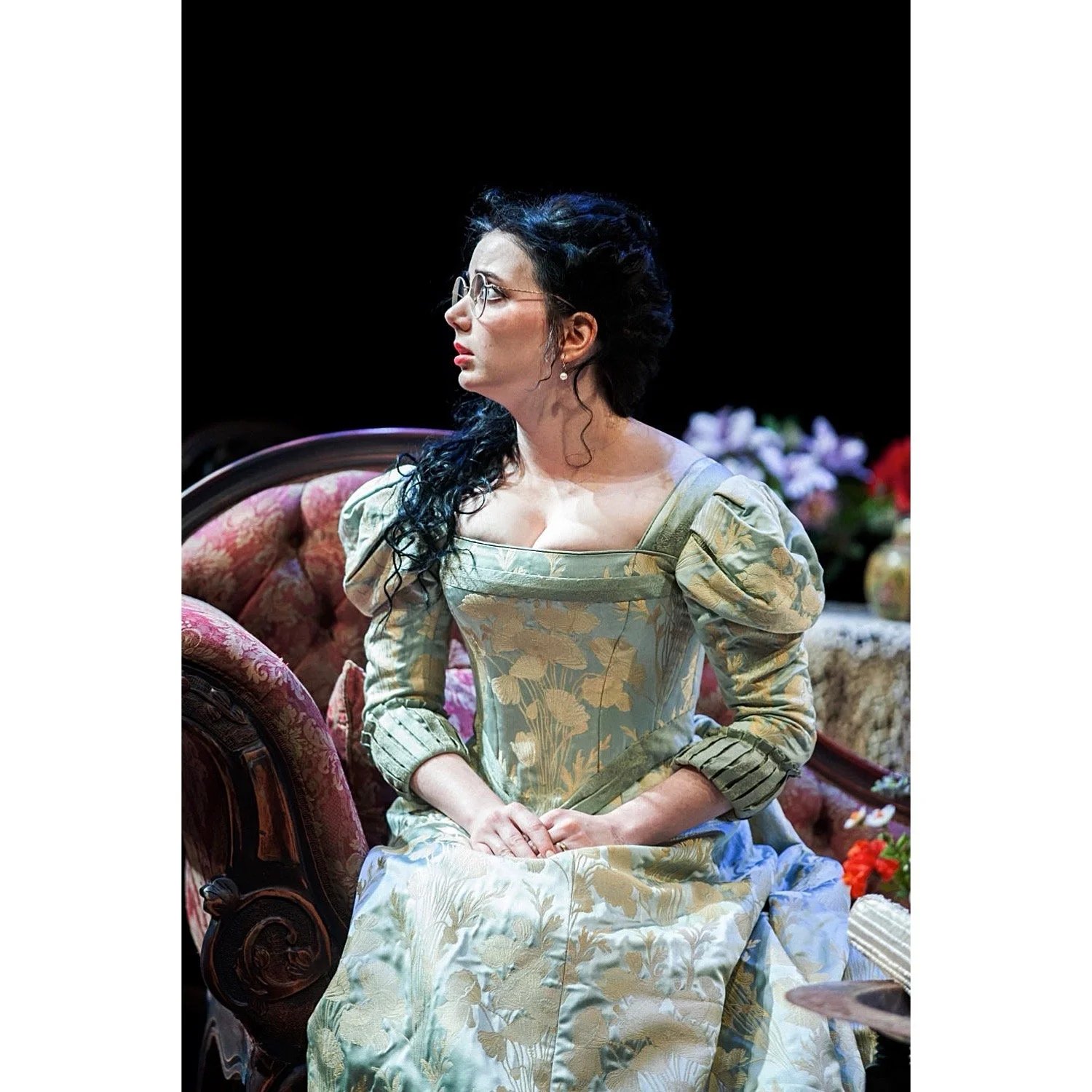 A woman with dark curly hair, glasses, and pearl earrings sits on an upholstered chair, wearing an 18th-century style green floral gown with puffed sleeves, in a theatrical scene with flowers in the background.