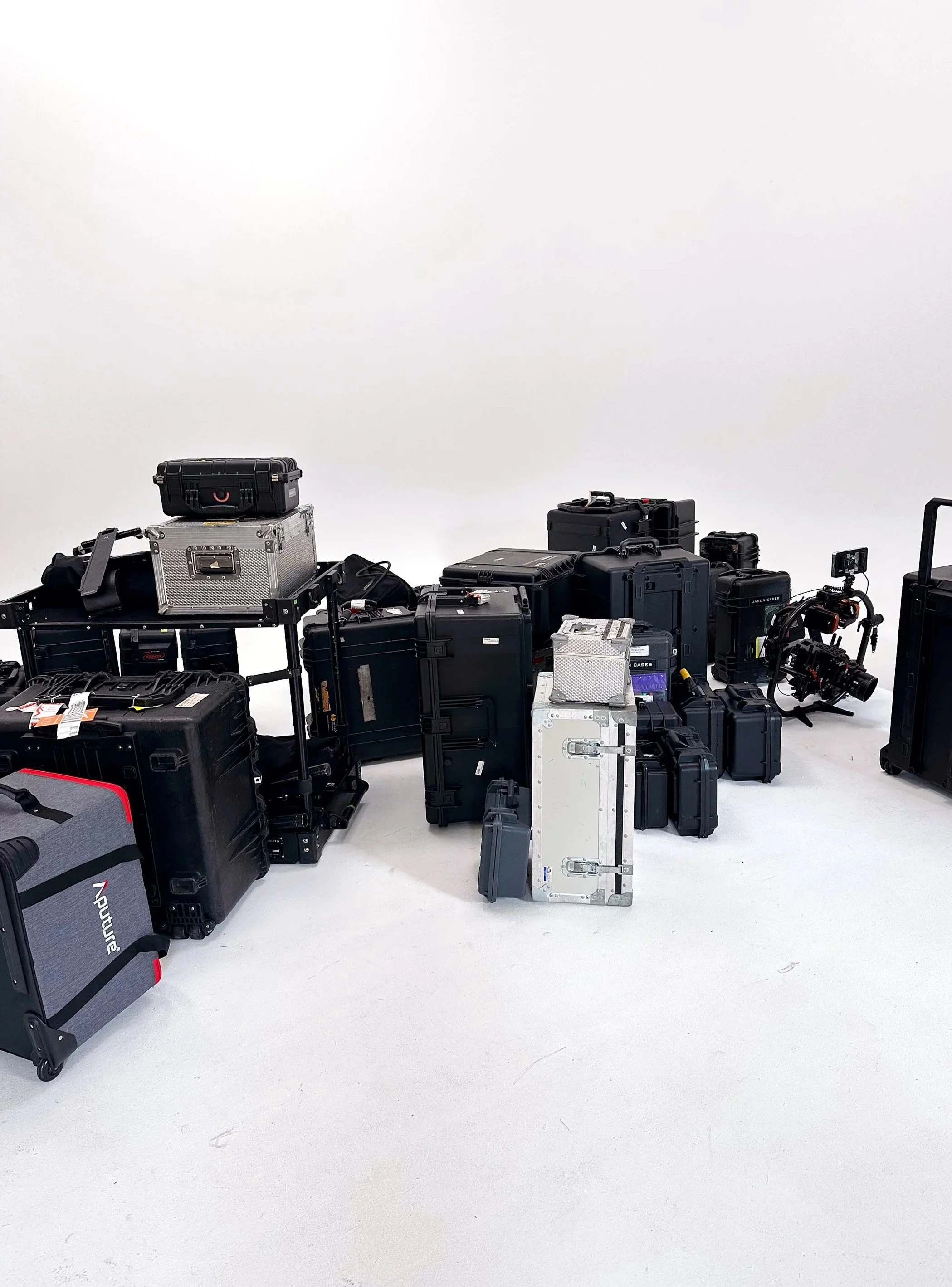 Various black, gray, and silver luggage cases and equipment scattered on a white floor against a white background.