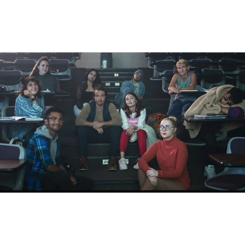 Group of nine students sitting in a classroom or lecture hall, some on stairs and some at desks, with notebooks and backpacks, in a casual and attentive pose.