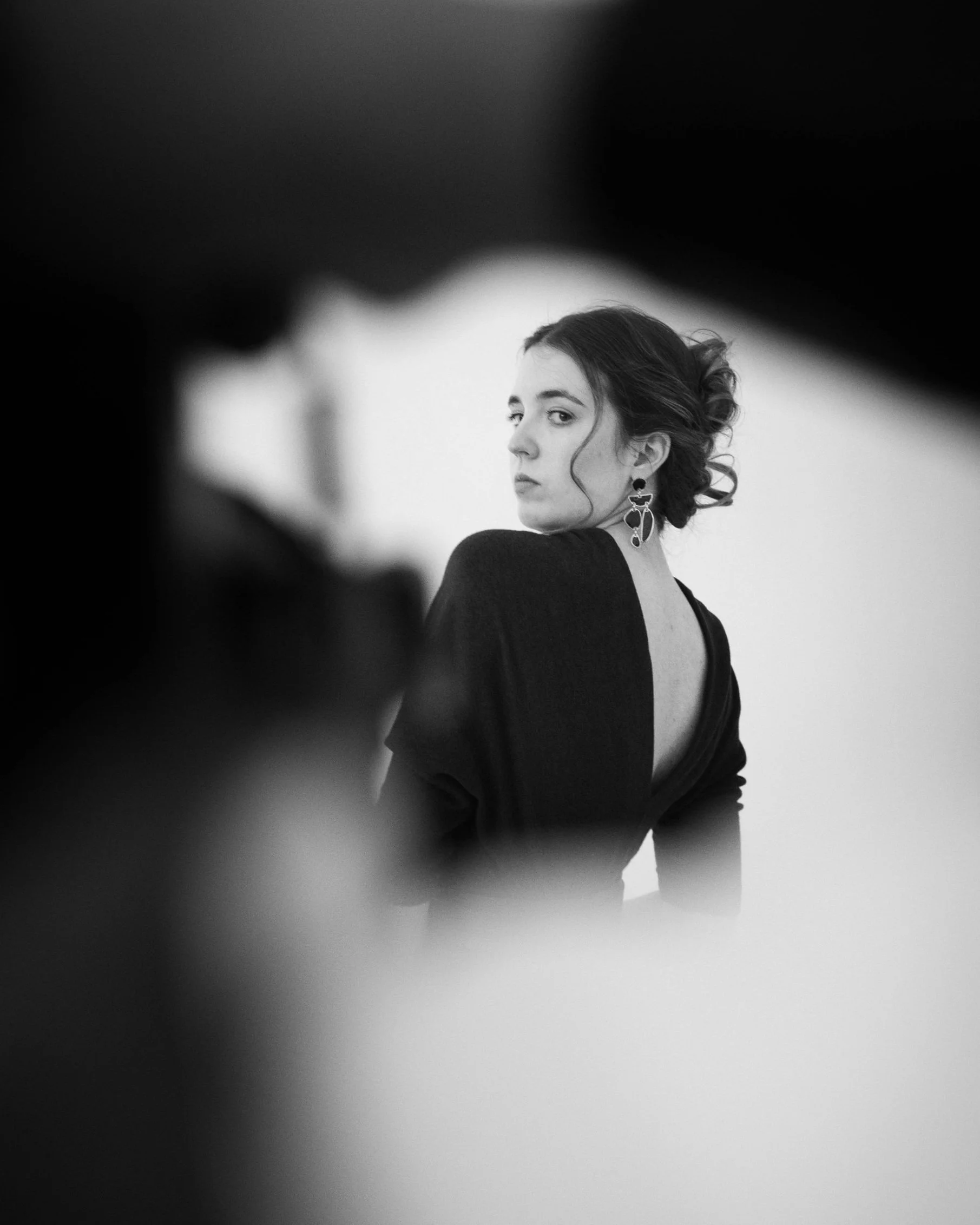 A woman with dark curly hair in an updo, wearing large earrings and a backless black dress, looks over her shoulder in a black-and-white portrait.