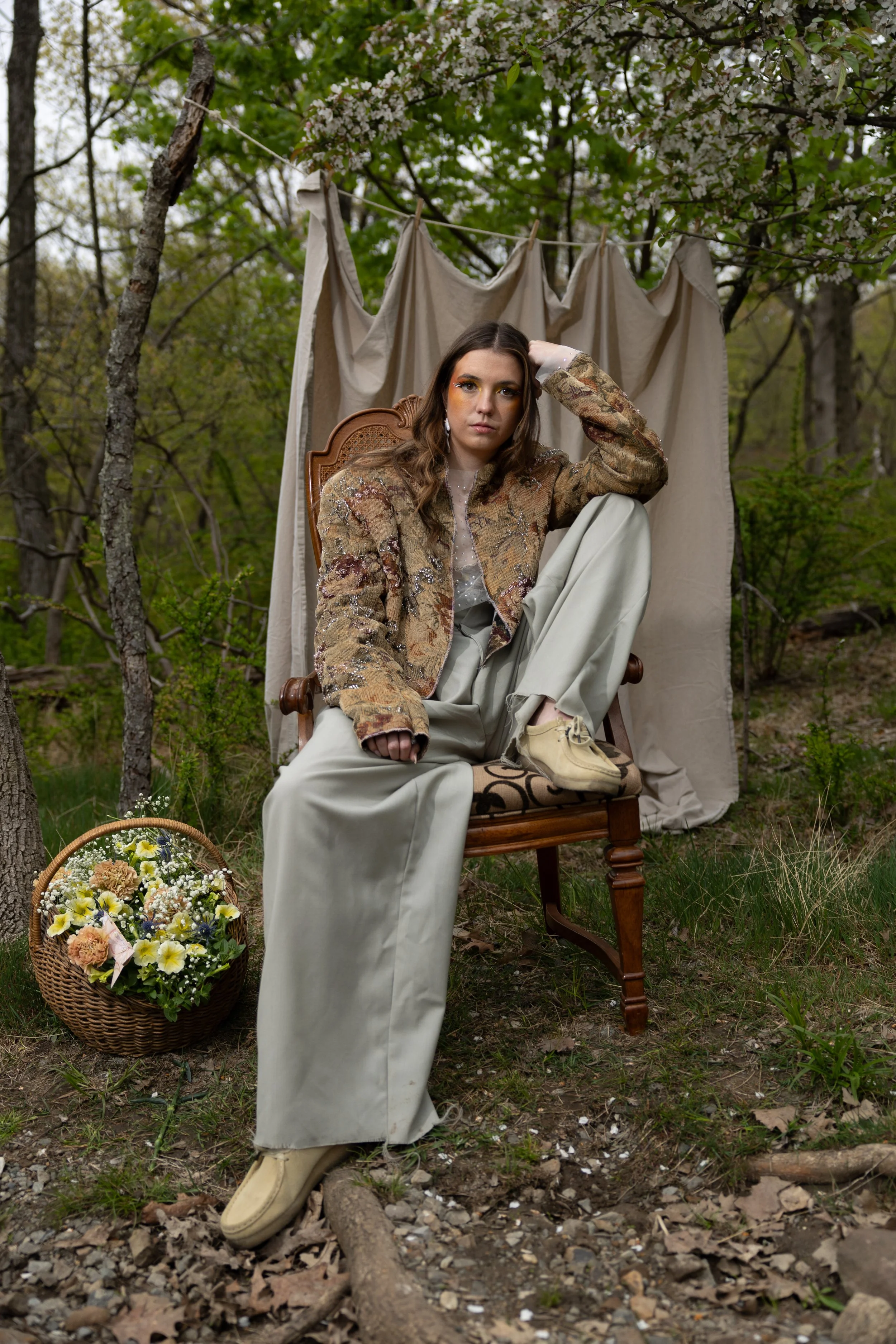 Young woman sitting on a vintage wooden chair outdoors, surrounded by trees and greenery, with a basket of flowers on the ground beside her. She wears a jacket, loose pants, and beige shoes, with a bathtub backdrop and a curtain hanging behind her.