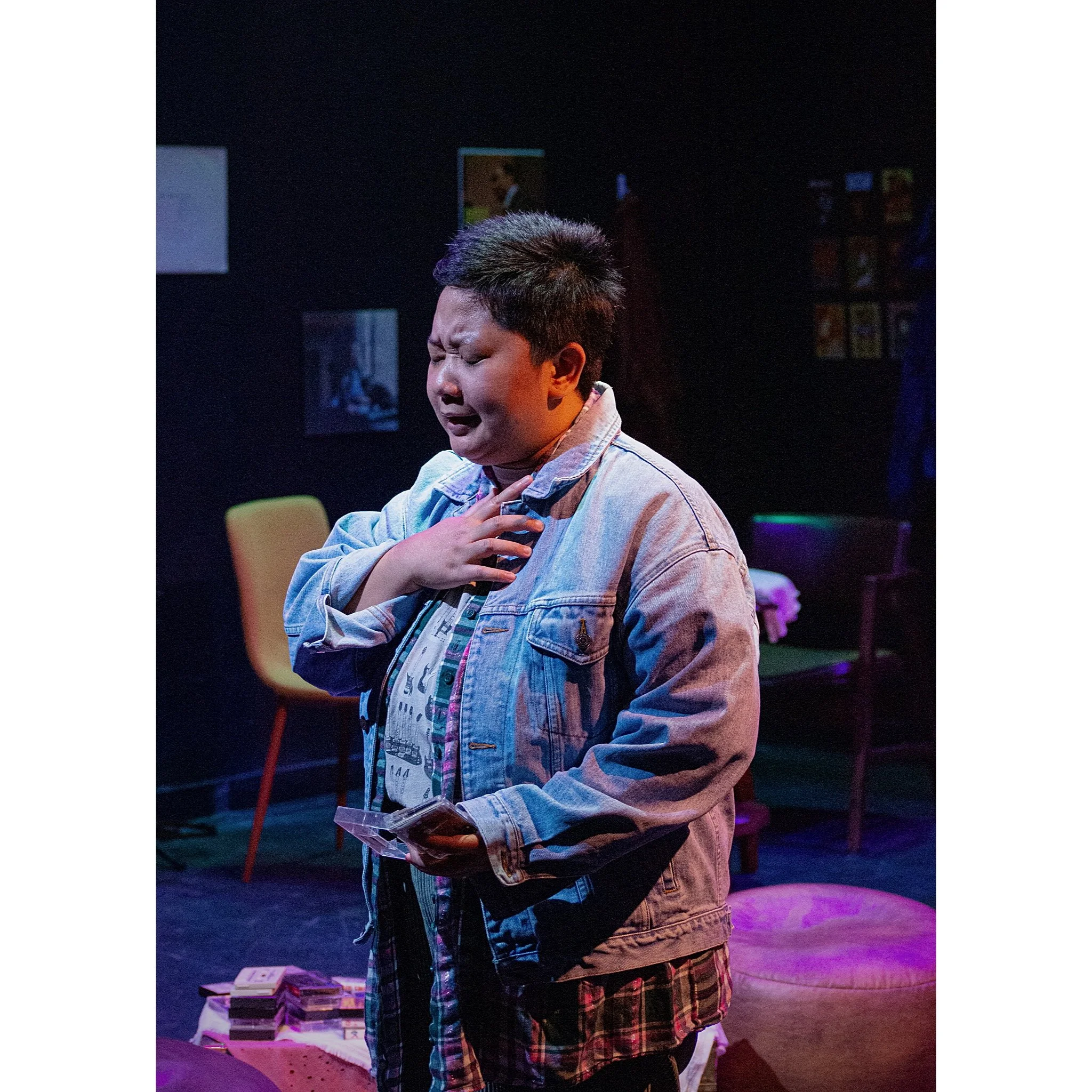A person with short dark hair wearing a denim jacket and plaid pants appears to be crying, with one hand on their chest and the other holding a phone, in a dimly lit room with colorful lighting and furniture.