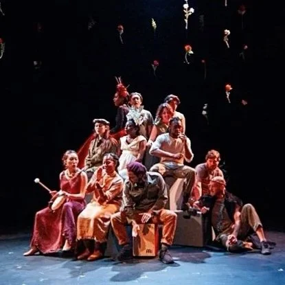 A group of performers dressed in period costumes on stage, with some sitting and some standing, surrounded by hanging flowers or decorations.