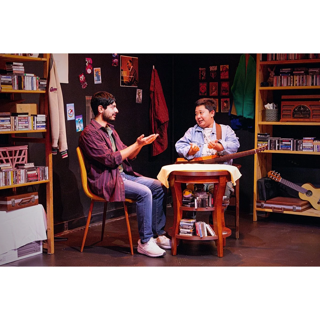 Two men having a conversation in a room filled with music and books, one of whom is holding a guitar, while the other gestures with his hands.