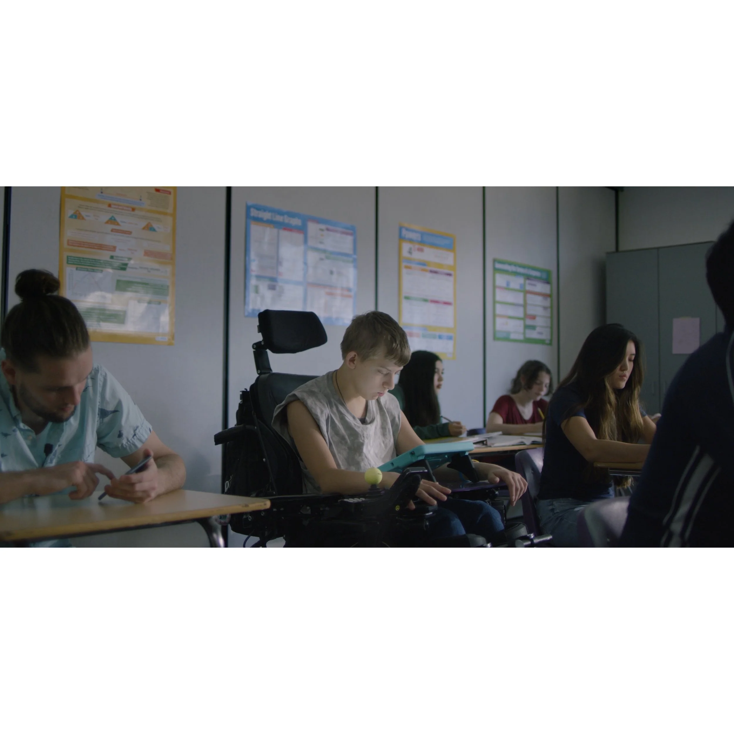 Students in classroom, one with a disability in a powered wheelchair, activities include using their phone, reading, and writing, classroom posters on the wall.