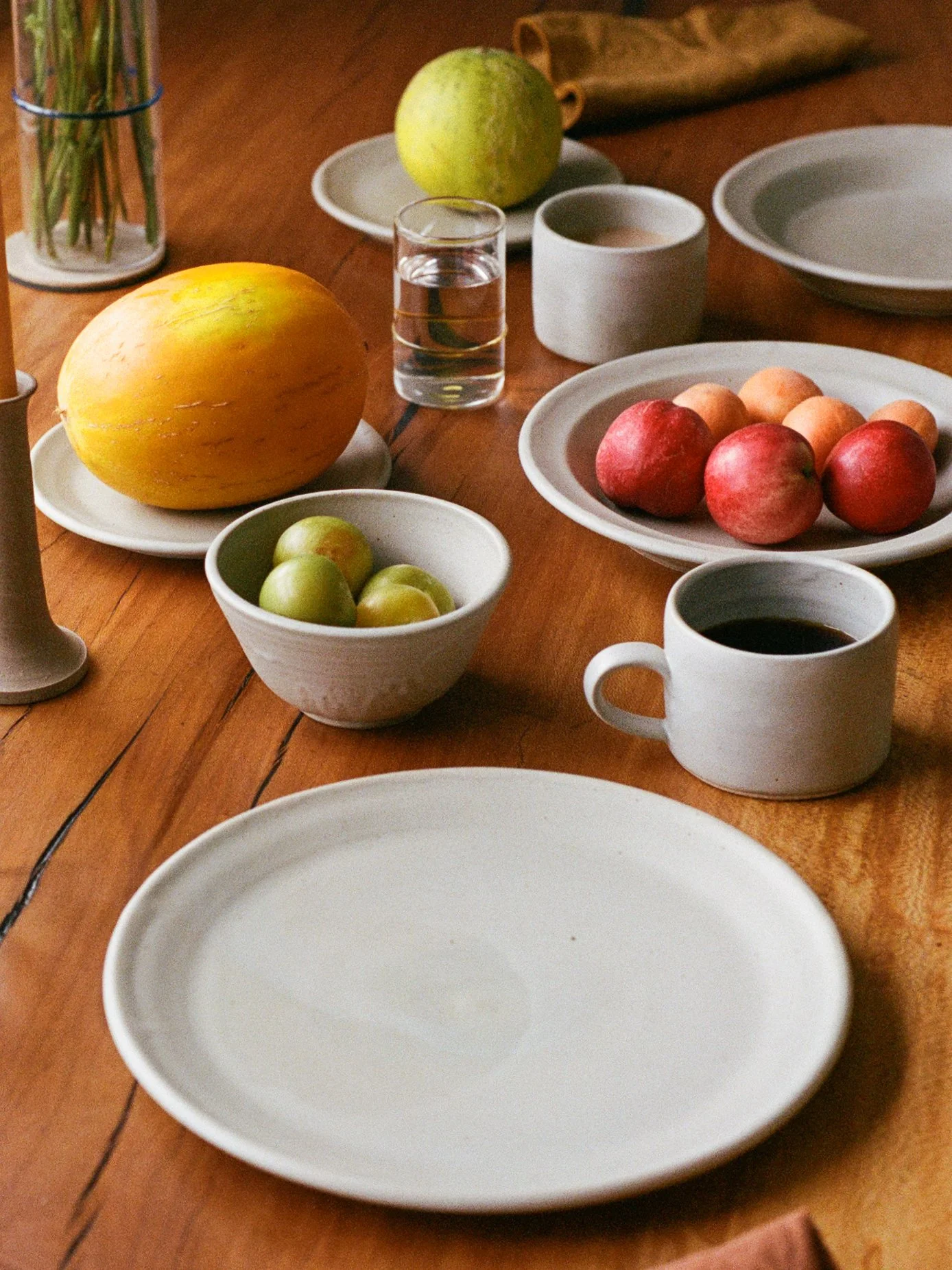 A wooden table set with bowls of green apples, peaches, and a large yellow melon, a glass of water, a cup of coffee, and empty gray plates and bowls, with a candle and a small green apple in the background.