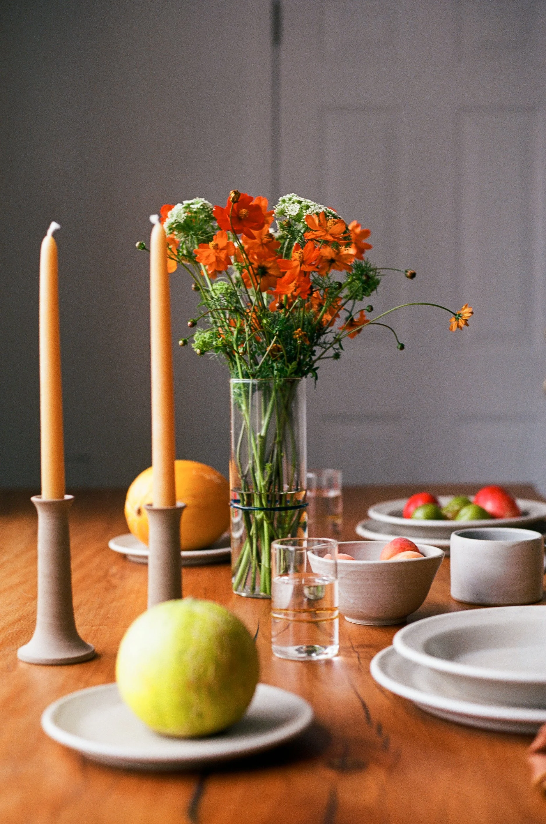 A wooden table set with white dishes, glass cups, a vase of orange and white flowers, and orange candles, with green apples and a pumpkin in the background.