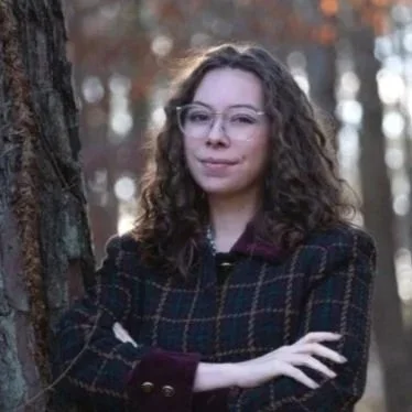 A white woman in a dark, collard coat leans up against a tree with her arms crossed, right over left, while looking directly into the camera and smiling with a small, closed-lipped smile. The woman has curly brown hair & wears wire-rimmed glasses.