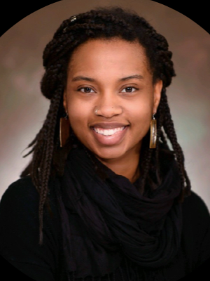 Black woman with dark braids, large dangly earrings, and a silver nose ring looks directly into the camera smiling with her top row of teeth showing.