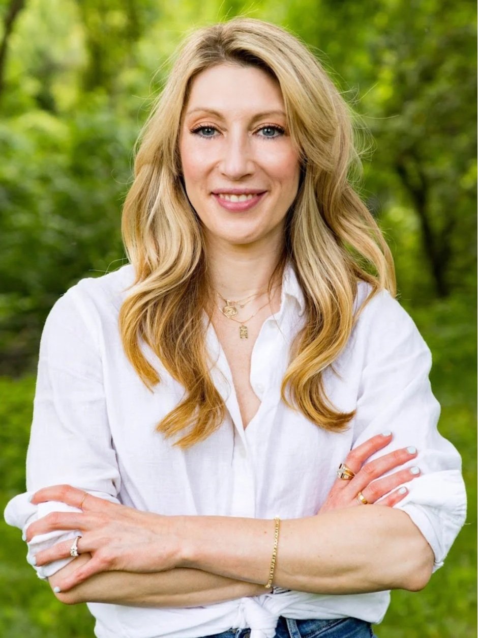 A white wommon with blonde brown hair and blue eyes wears a white collared shirt tucked into jeans and several necklaces, rings, and bracelets stands with arms crossed looking directly into the camera and smiles a crooked smile.
