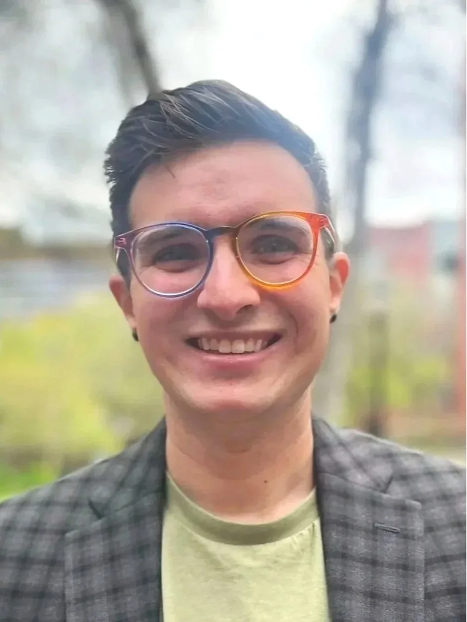 Young white man in a blazer with ear piercings and rainbow glasses smiles directly into the camera.