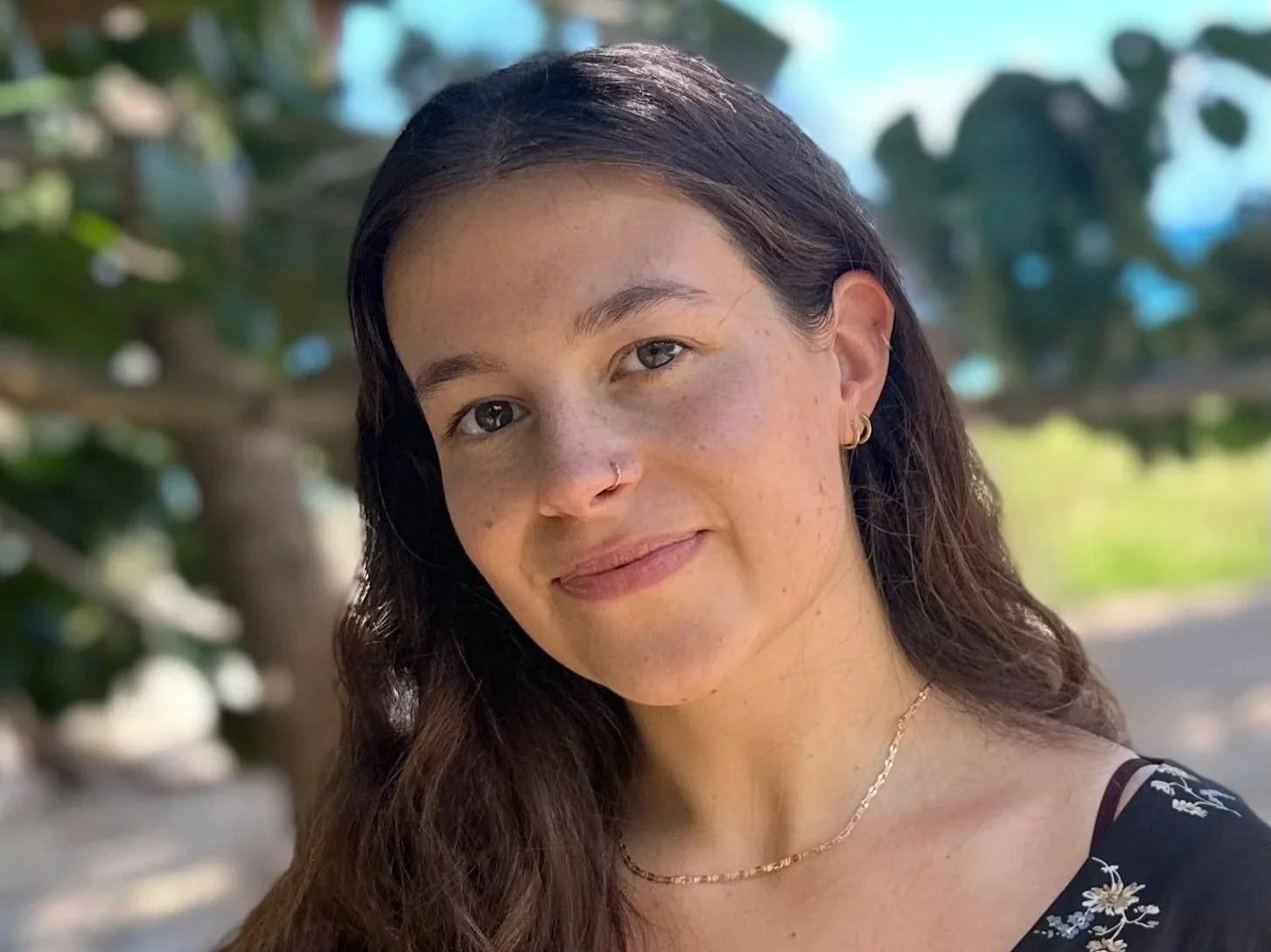 A white woman with dark brown shoulder-length hair worn lose, brown eyes, freckles, with no makeup, a nose ring, three earring hoops, and a gold necklace smiles a faint, closed-lipped smile while looking directly at the camera.