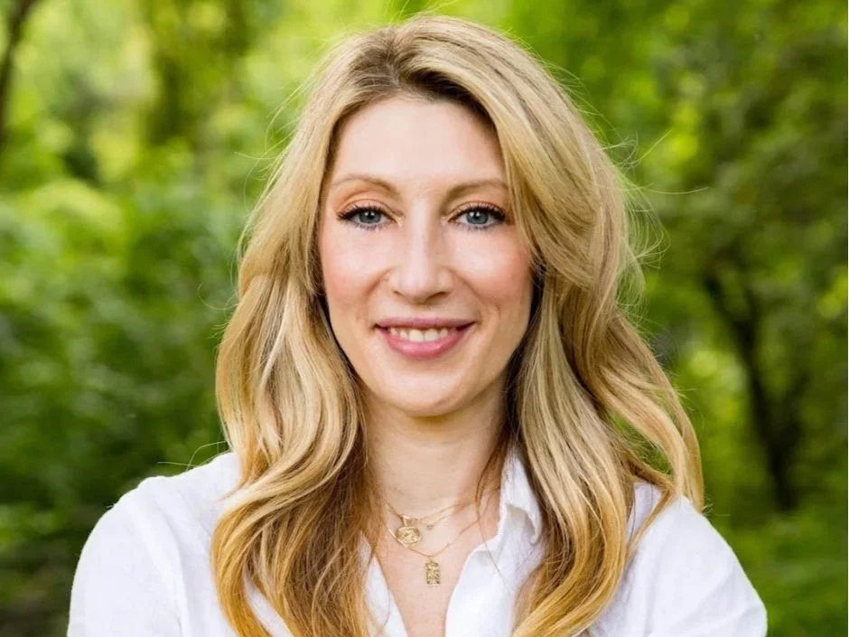 A white wommon with blonde brown hair, blue eyes, and light makeup wears a white collared shirt and several gold necklaces looks directly into the camera and smiles a crooked grin.