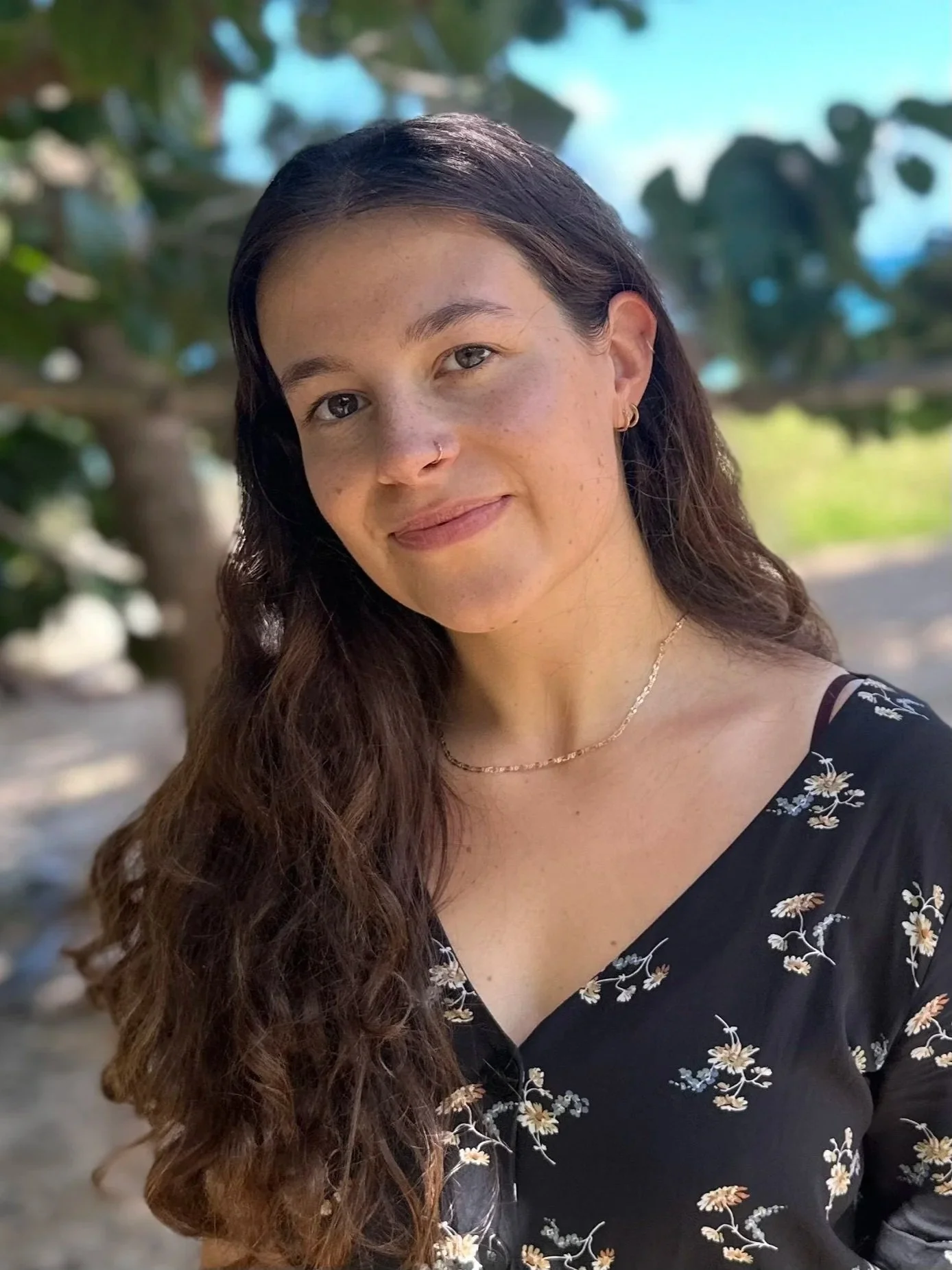 A white woman with dark brown shoulder-length hair worn lose, brown eyes, freckles, with no makeup, a nose ring, three earring hoops, and a gold necklace smiles a faint, closed-lipped smile while looking directly at the camera.