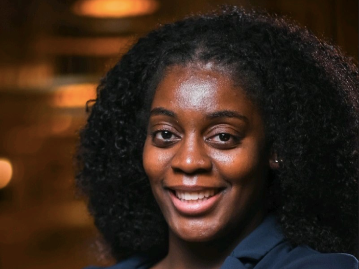 A Black wommon with curly black hair and dark brown eyes looks directly at the camera and smiles a large, toothy grin.
