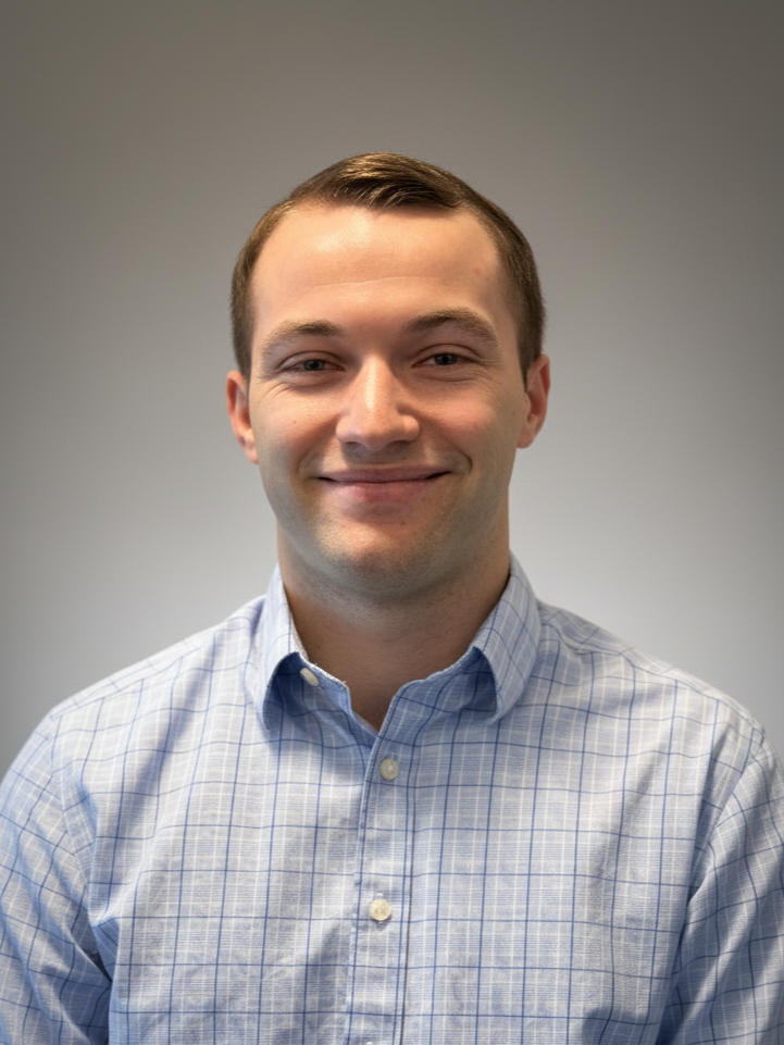 Young white man with brown slicked and combed over hair wearing a blue checkered dress shirt looks into the camera and smiles closed lipped.
