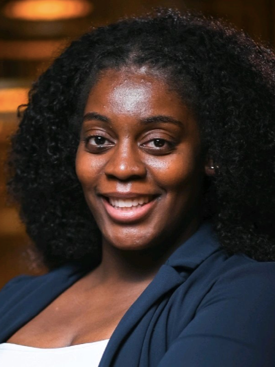 A Black wommon with curly black hair and dark brown eyes wearing a white top covered by a blue blazer and minimal makeup looks directly at the camera and smiles a large, toothy grin.