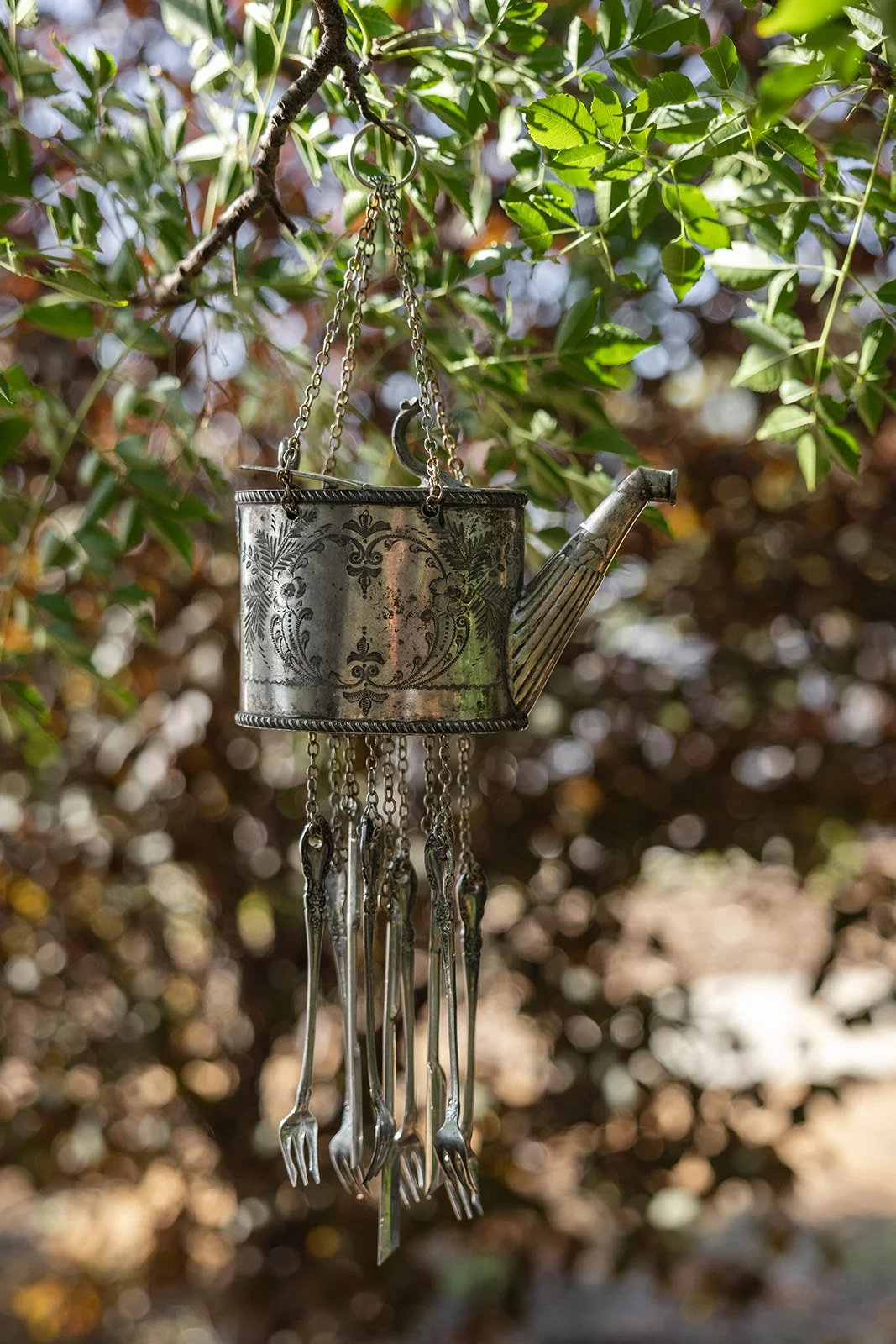 A decorative metal watering can hanging from a tree branch with green leaves. The watering can has intricate engraved patterns and chains hanging from its spout, ending in miniature fork and spoon charms.