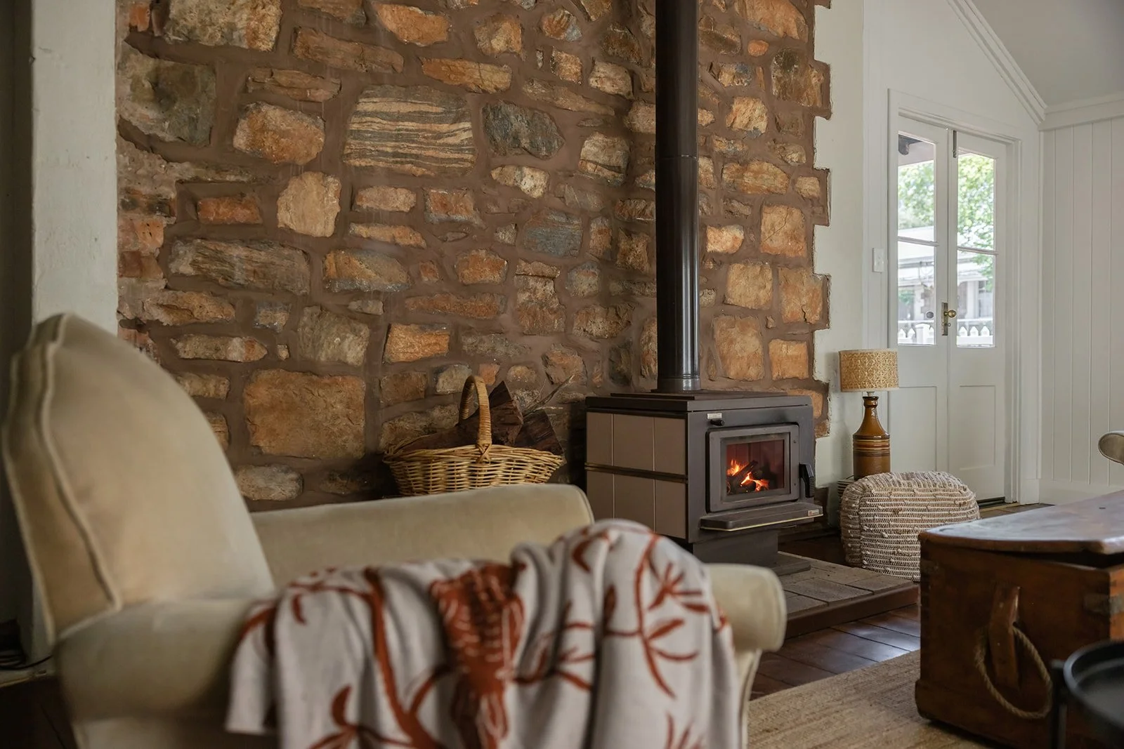 Living room with a stone fireplace, beige armchair with a patterned throw, wicker basket, and a wood stove with a fire, next to a white door with windows and a small side table with a lamp.