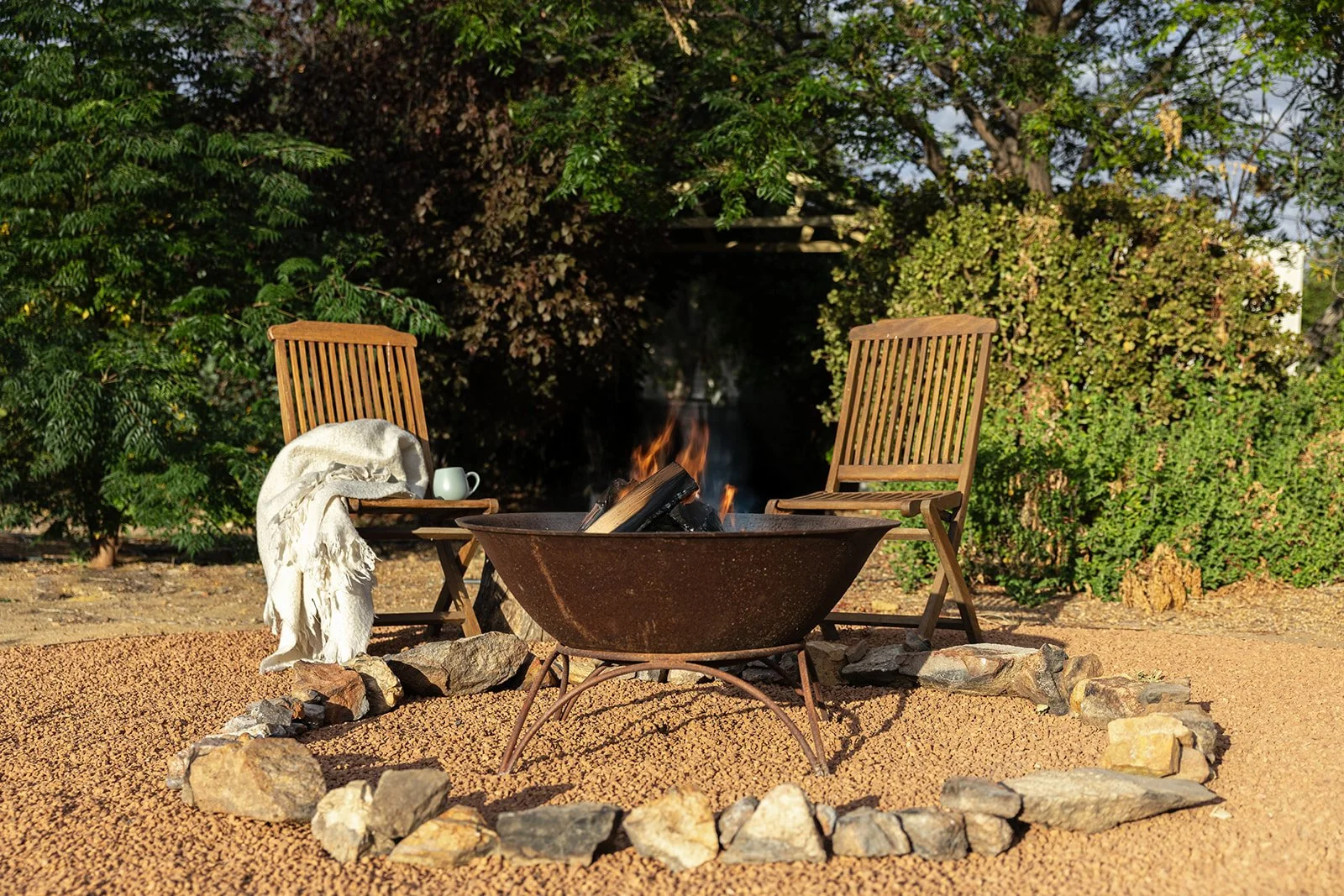 Two wooden chairs placed around a fire pit with a burning fire, a white blanket draped over one chair, and a white mug on the chair's armrest, set outdoors with green bushes and trees in the background.
