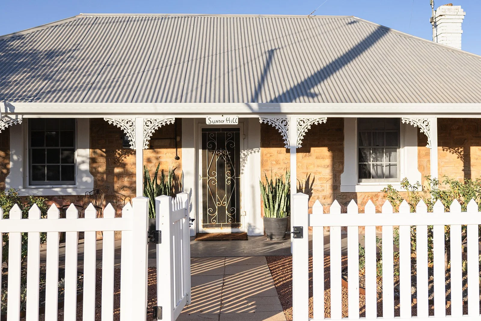Front verandah of Sunny Hill, a heritage listed stone cottage in York, WA with a metal roof, white decorative trim, a white picket fence, and a front garden with potted plants and shrubs.