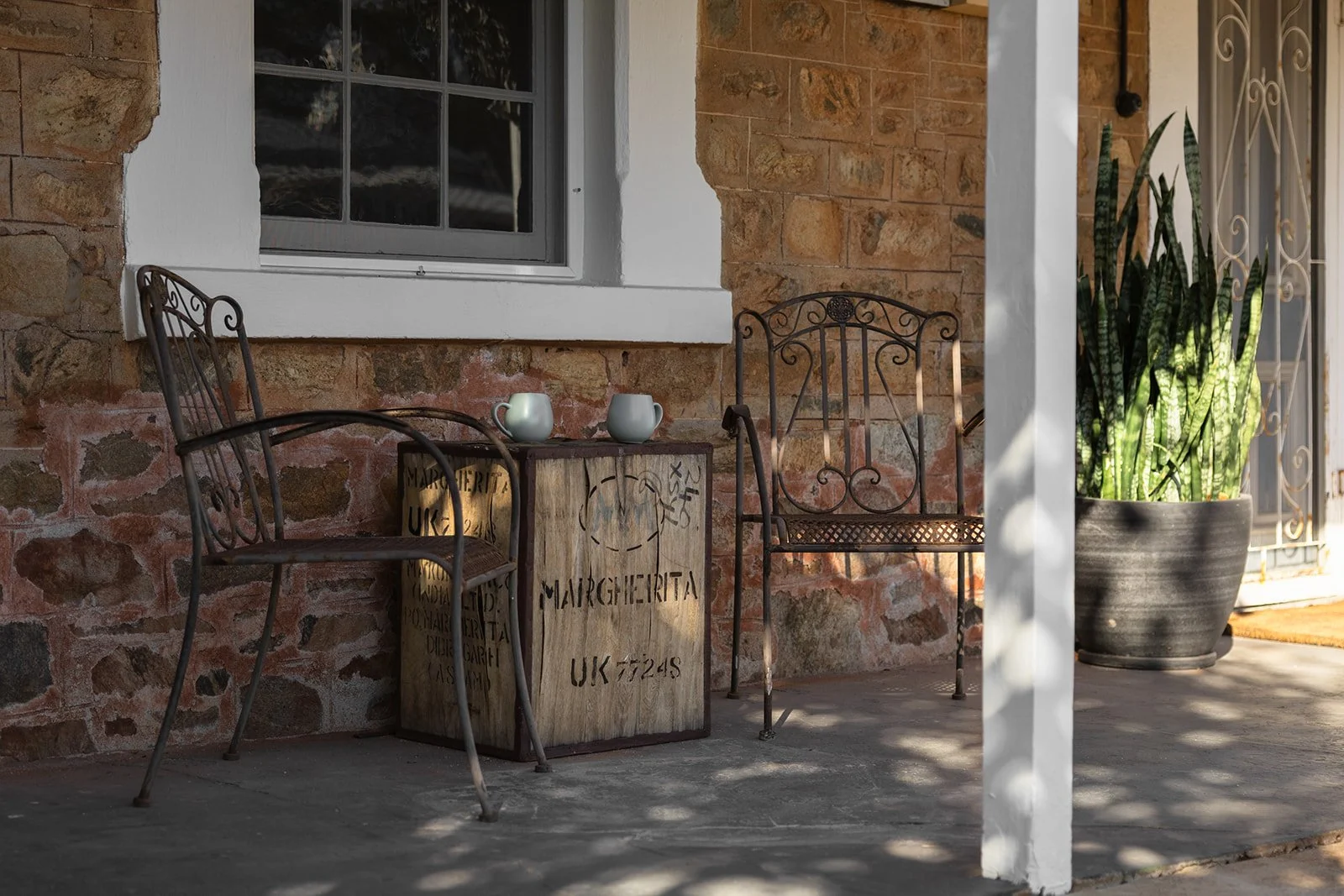 Patio with two metal chairs, a wooden crate with the word 'MARGHERITA' written on it, and a large potted snake plant in front of a brick wall with a window.