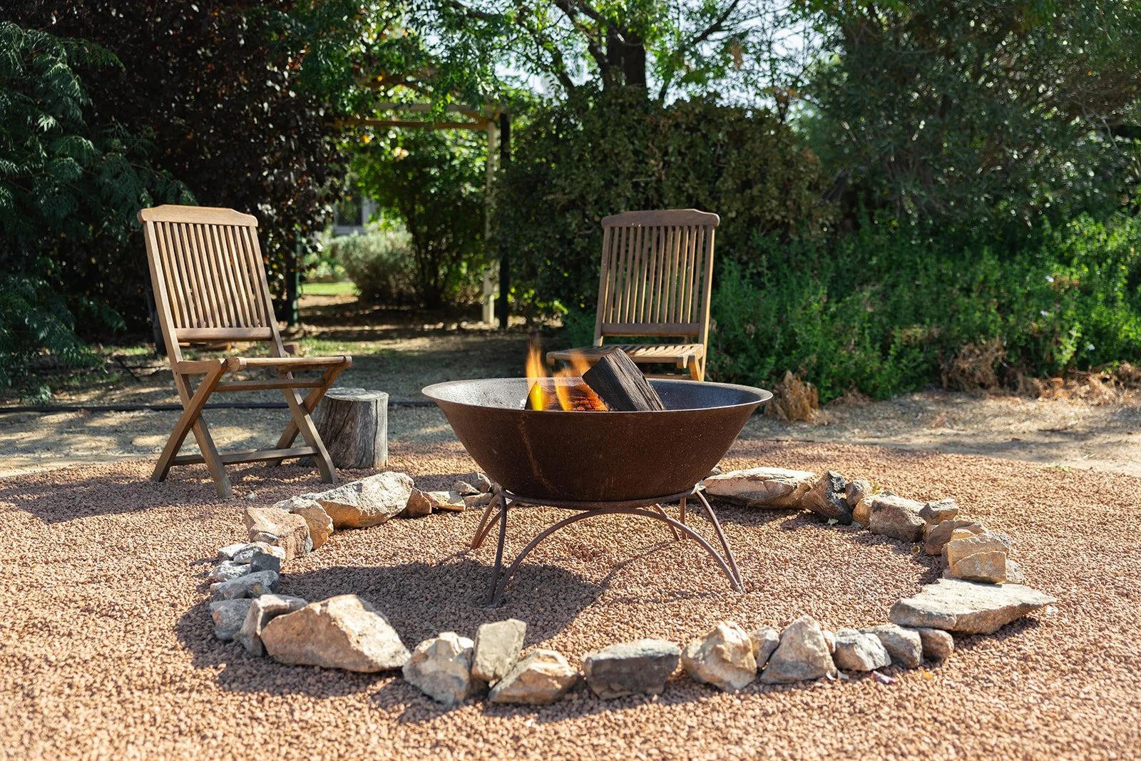 A backyard fire pit surrounded by rocks with two wooden chairs and a small wooden stump, with green bushes and trees in the background.