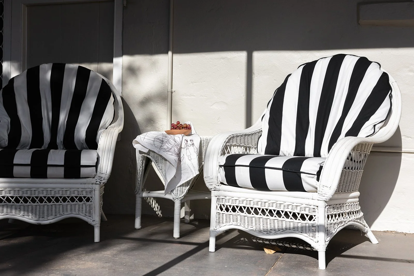 Two white wicker chairs with black and white striped cushions, next to a small white side table on the back verandah of an old farm house cottage in York, WA with sunlight and shadows.