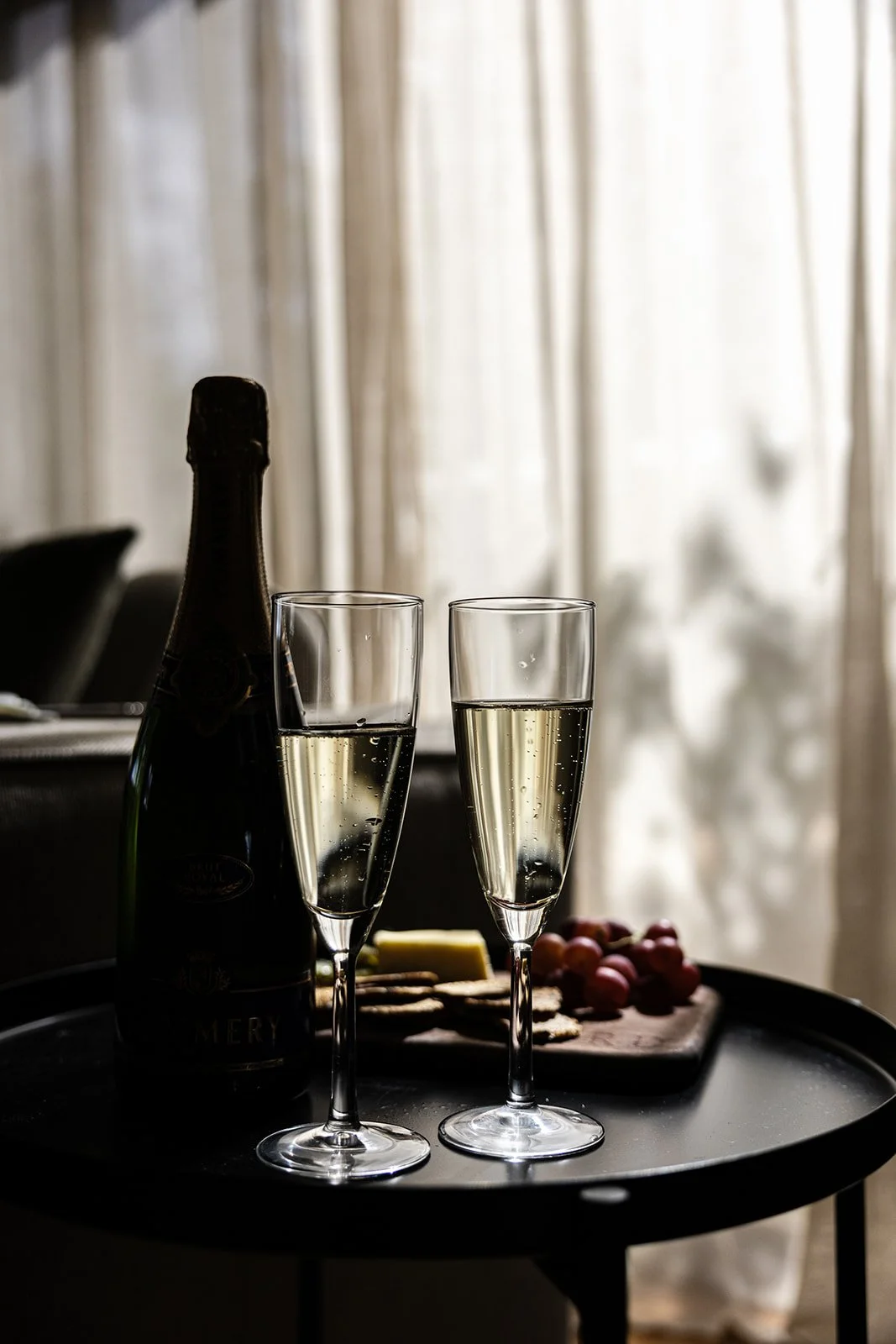 Two glasses of champagne with bubbles, a bottle of champagne, a cheese platter, grapes, and crackers on a round black tray, with a background of beige curtains.