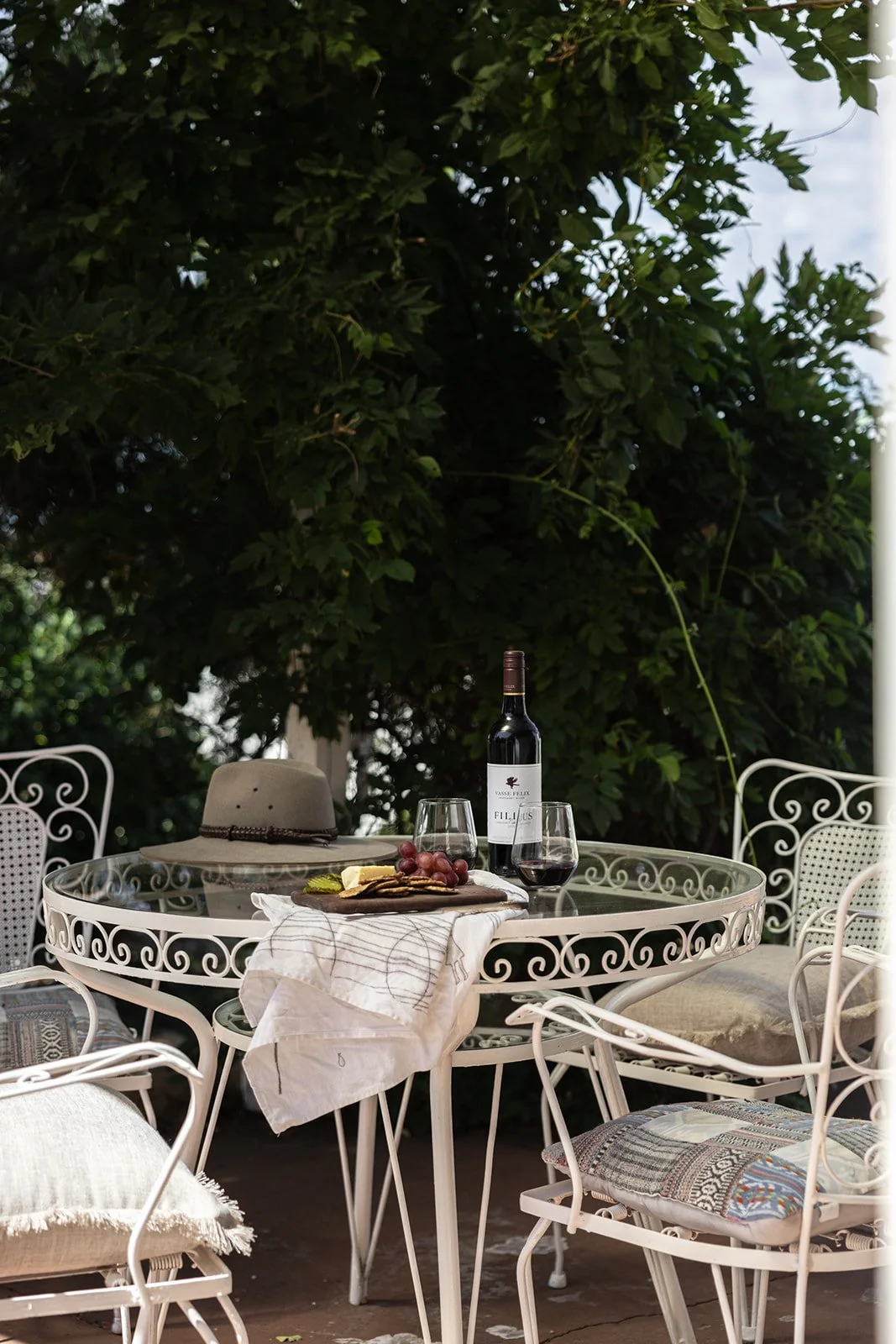 Outdoor table set with a bottle of red wine, two wine glasses, grapes, cheese, and crackers, surrounded by white wrought iron chairs and decorated cushions, under a large leafy tree.