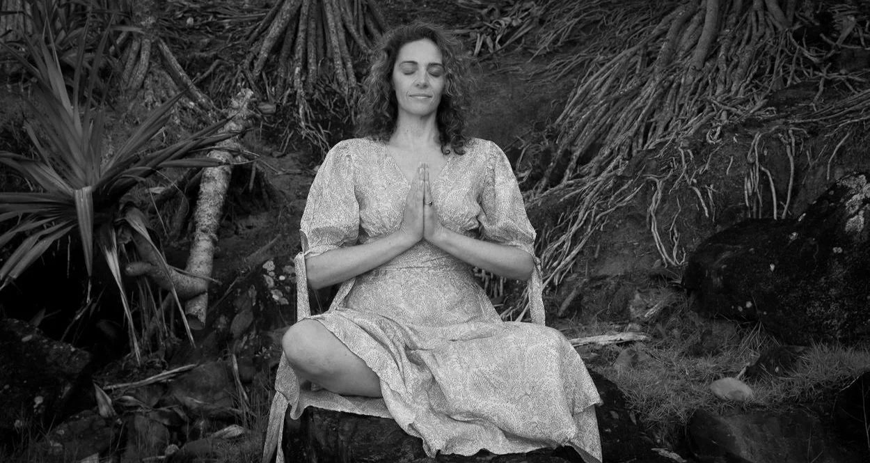 A woman meditating outdoors on a rock, sitting cross-legged with hands in prayer position, eyes closed, surrounded by natural foliage.