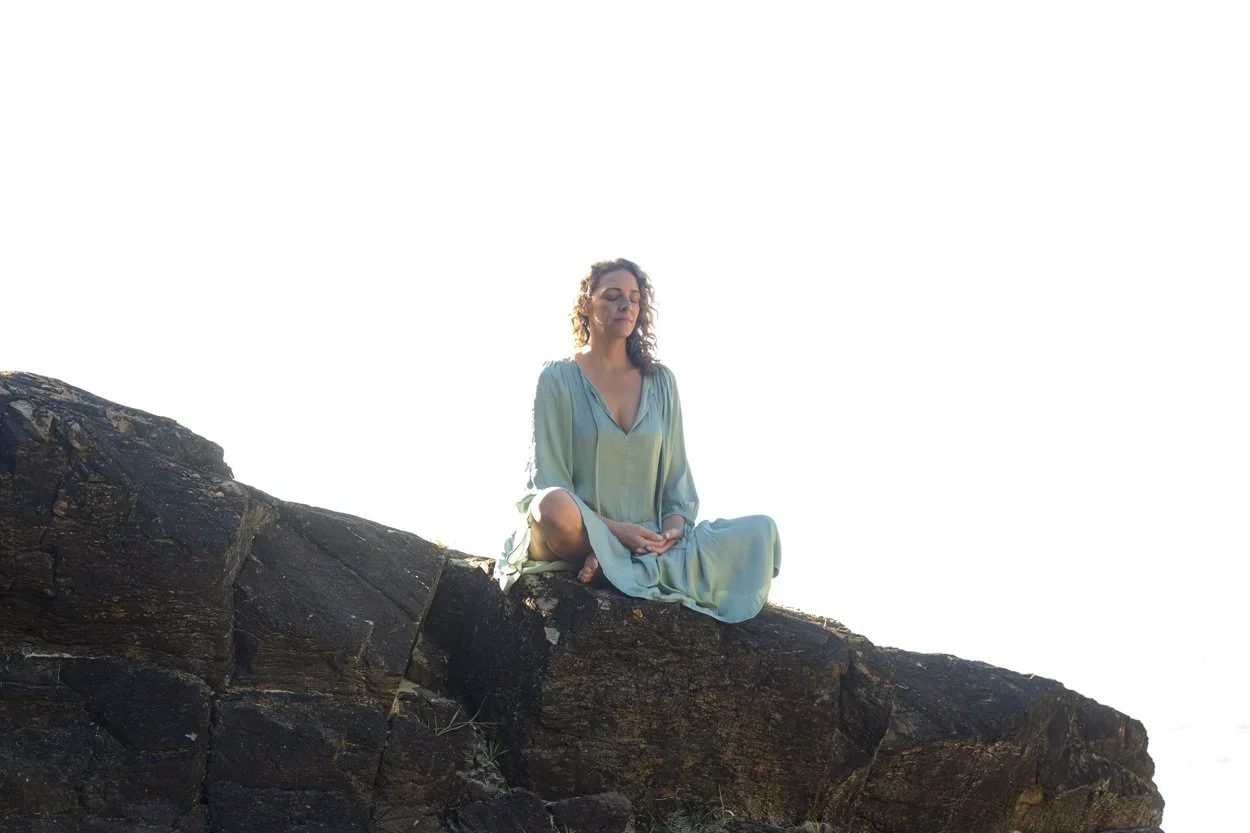 Woman in a light green dress practicing meditation while sitting cross-legged on a rocky outcrop outdoors during daytime.