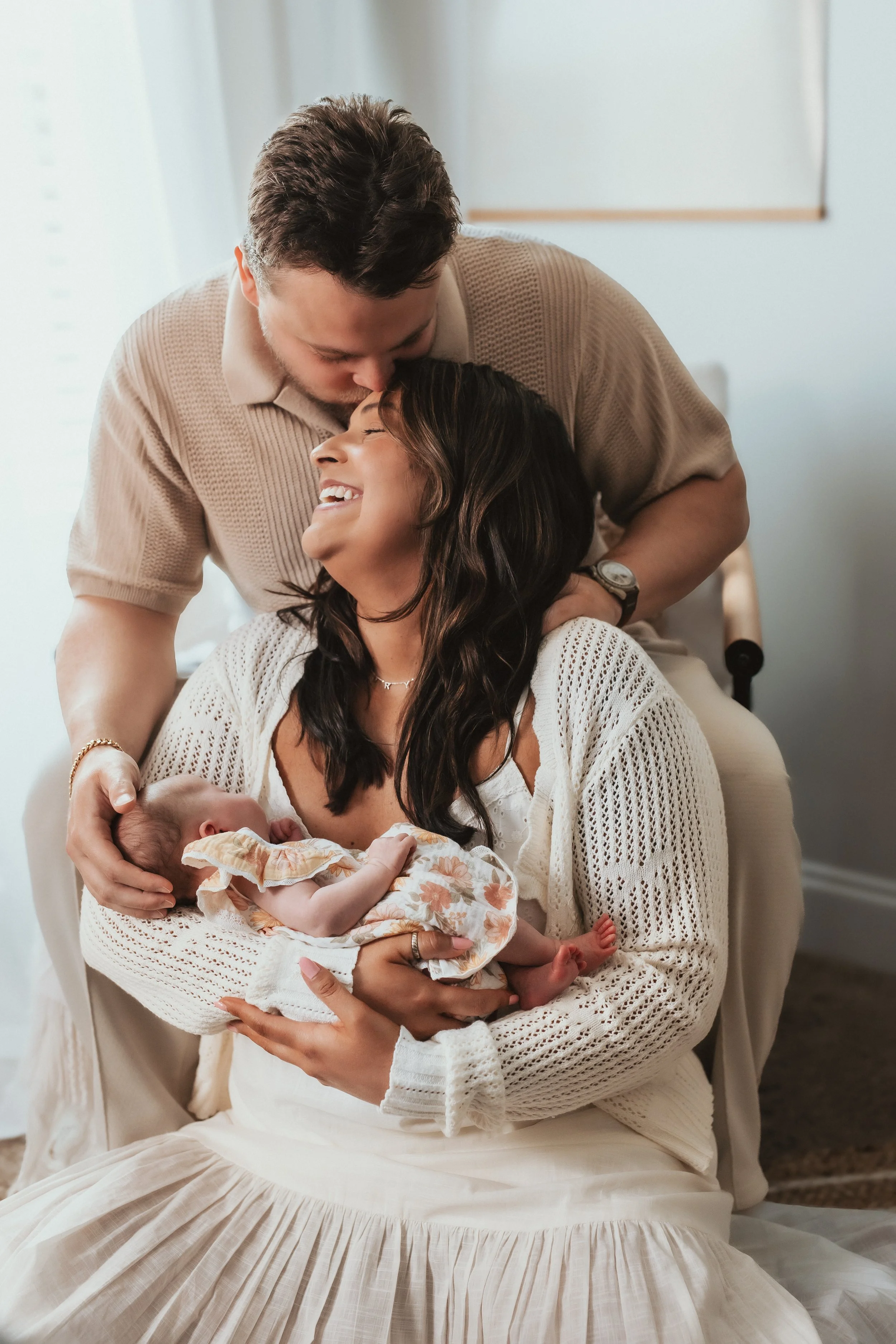 A woman holding a newborn baby in her arms, sitting on a hospital bed, with a man kissing her on the head, in a hospital room with light-colored walls.