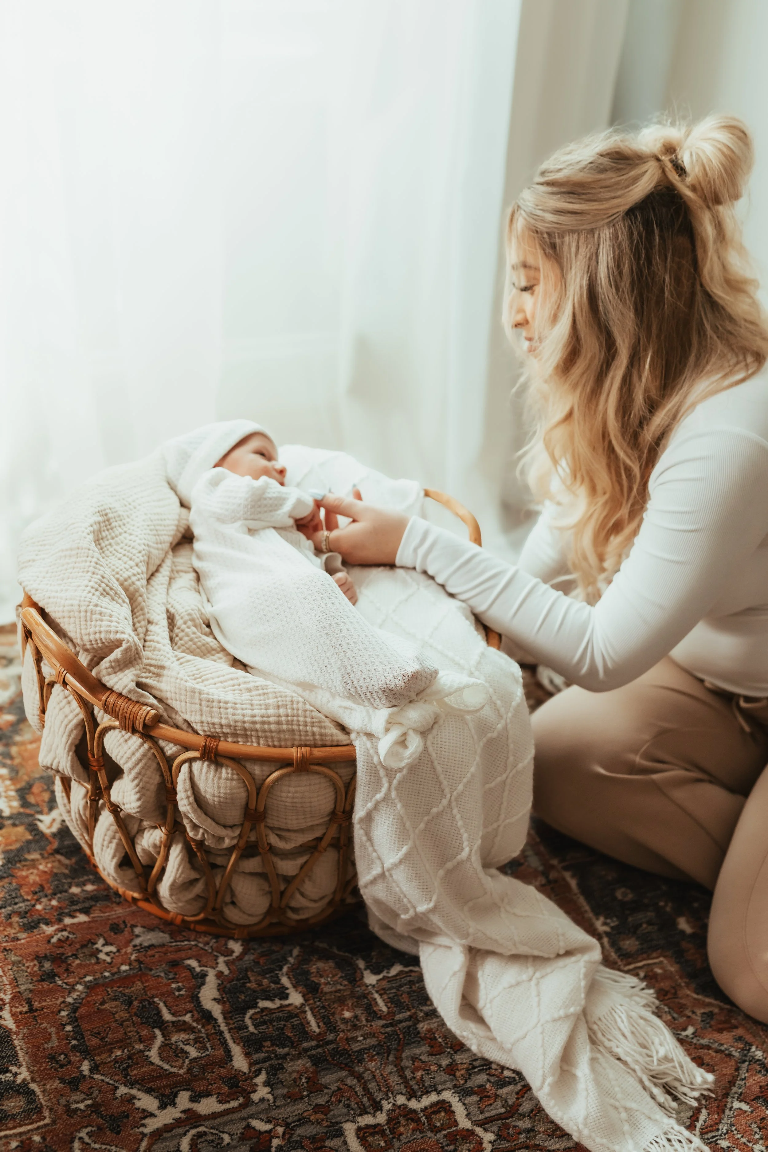 A woman with long blonde hair is kneeling beside a bassinet, gently touching a newborn baby who is lying inside. The baby is dressed in a white knit hat and a white outfit, wrapped in a cream blanket. The scene is softly lit by natural light.