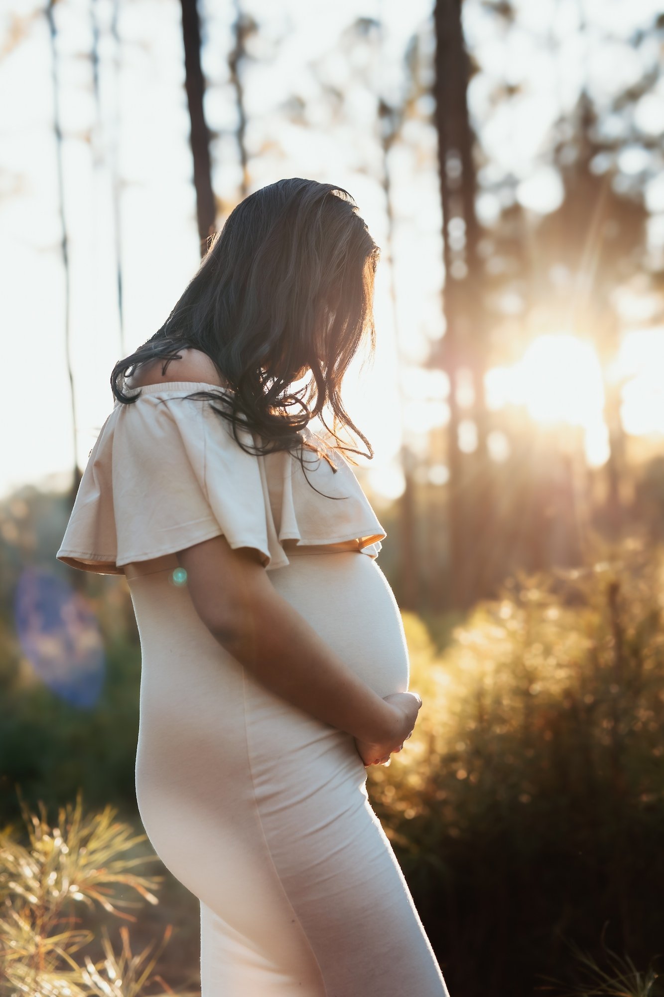 Pregnant woman in a white dress standing outdoors in a forest during sunset, gently holding her belly and looking downward.