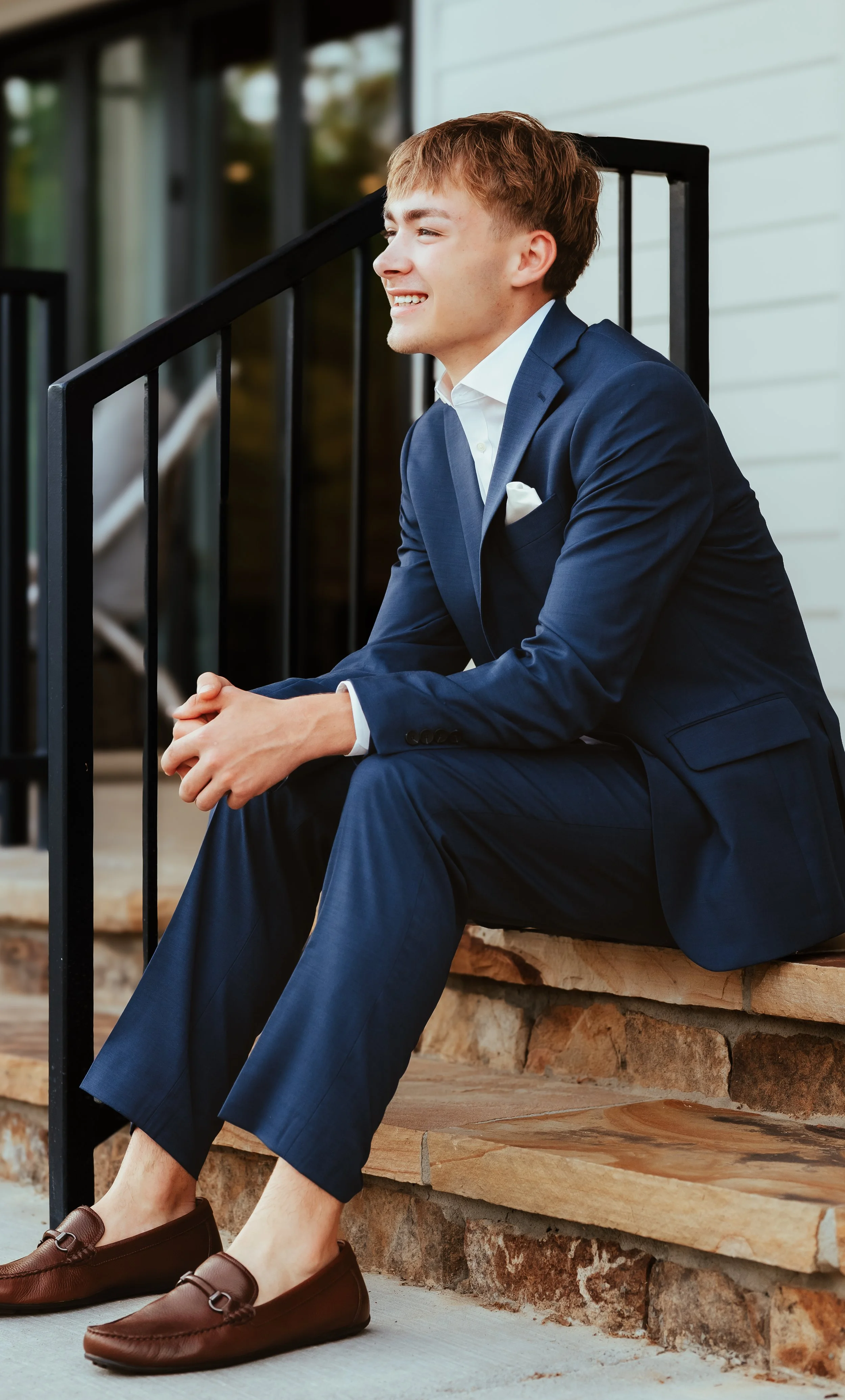 A young man in a blue suit sitting on stone steps outside, smiling and looking to the side, with a porch and glass doors in the background.