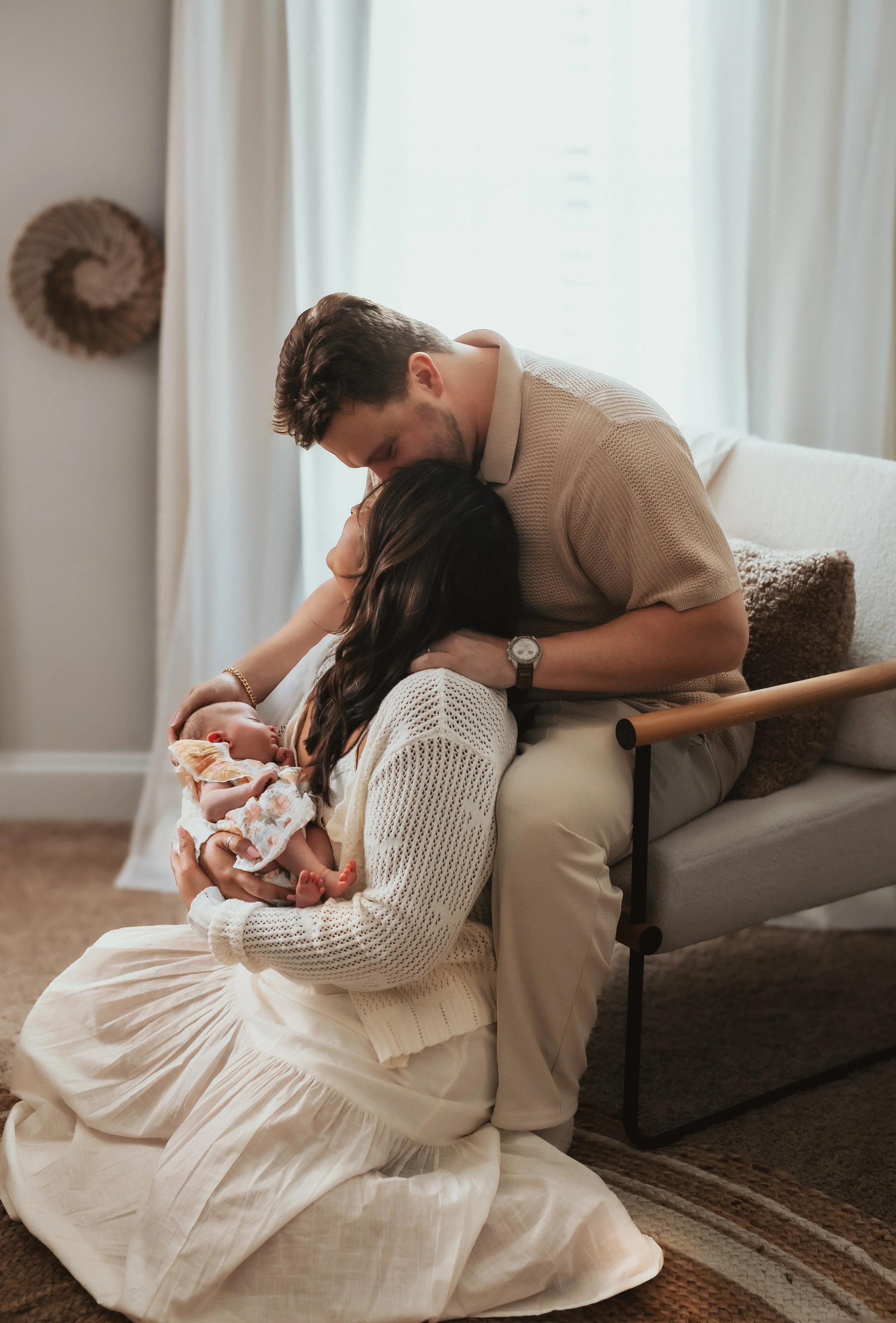 A family celebrating the birth of a newborn baby. The father is leaning over and kissing the mother on the forehead while she is holding their sleeping newborn girl in her arms. The scene is in a cozy, softly lit living room with curtains and decorat