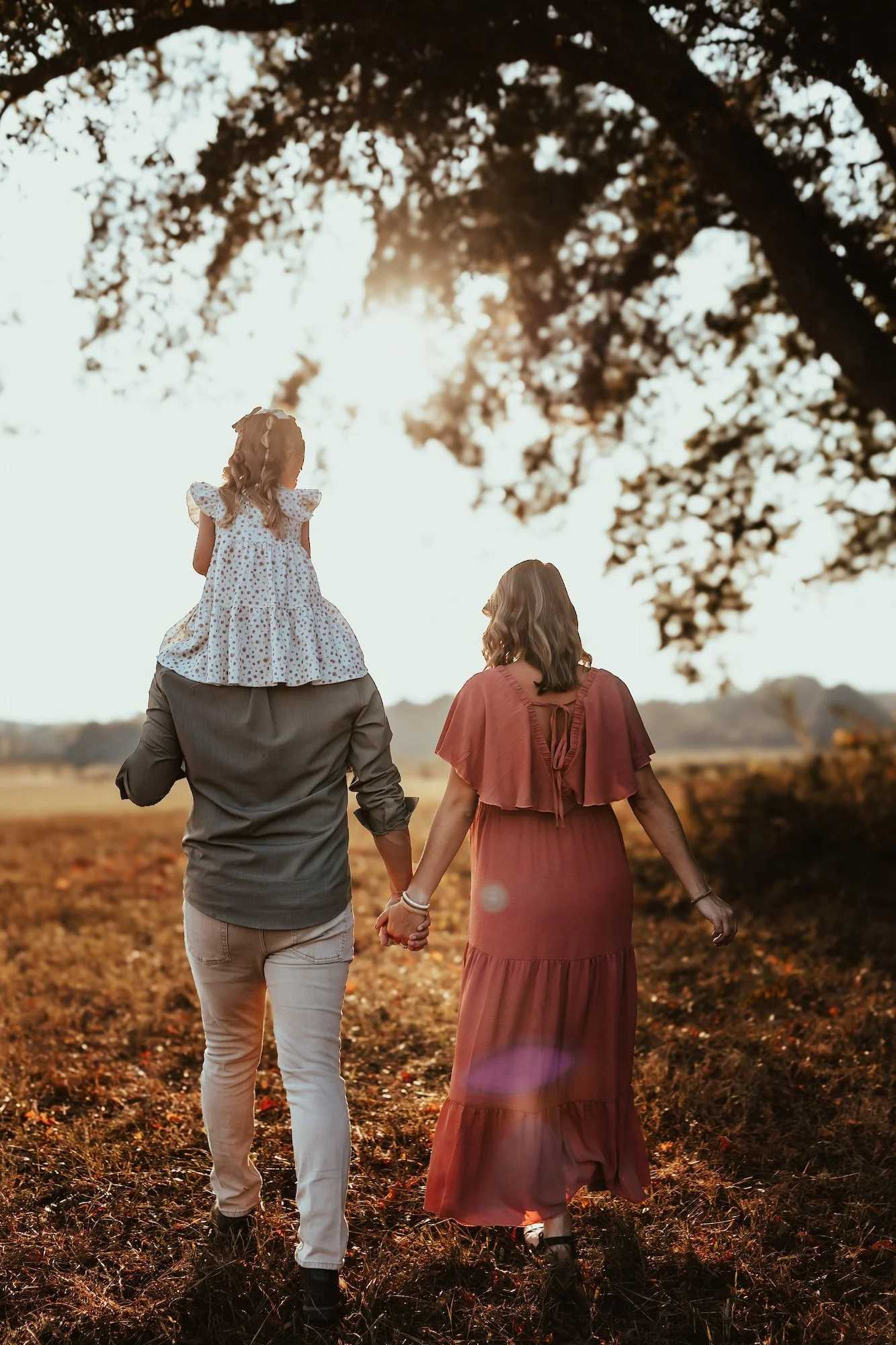 A family of three walking hand in hand through a field at sunset, with the father carrying a young girl on his shoulders and the mother holding his hand.