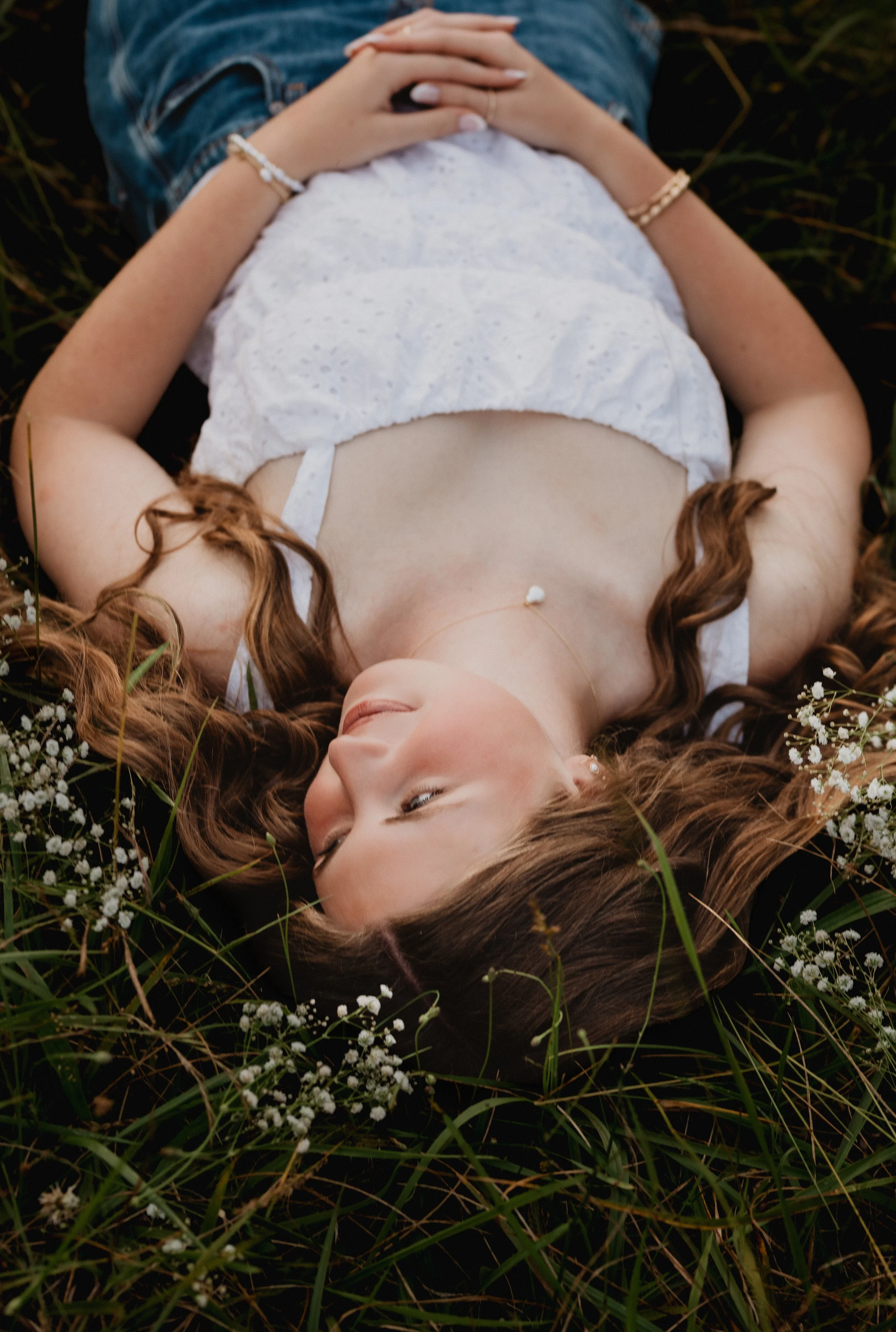 A woman with long brown hair lies on her back on grass with small white flowers, wearing a white dress and jewelry, with her eyes closed.