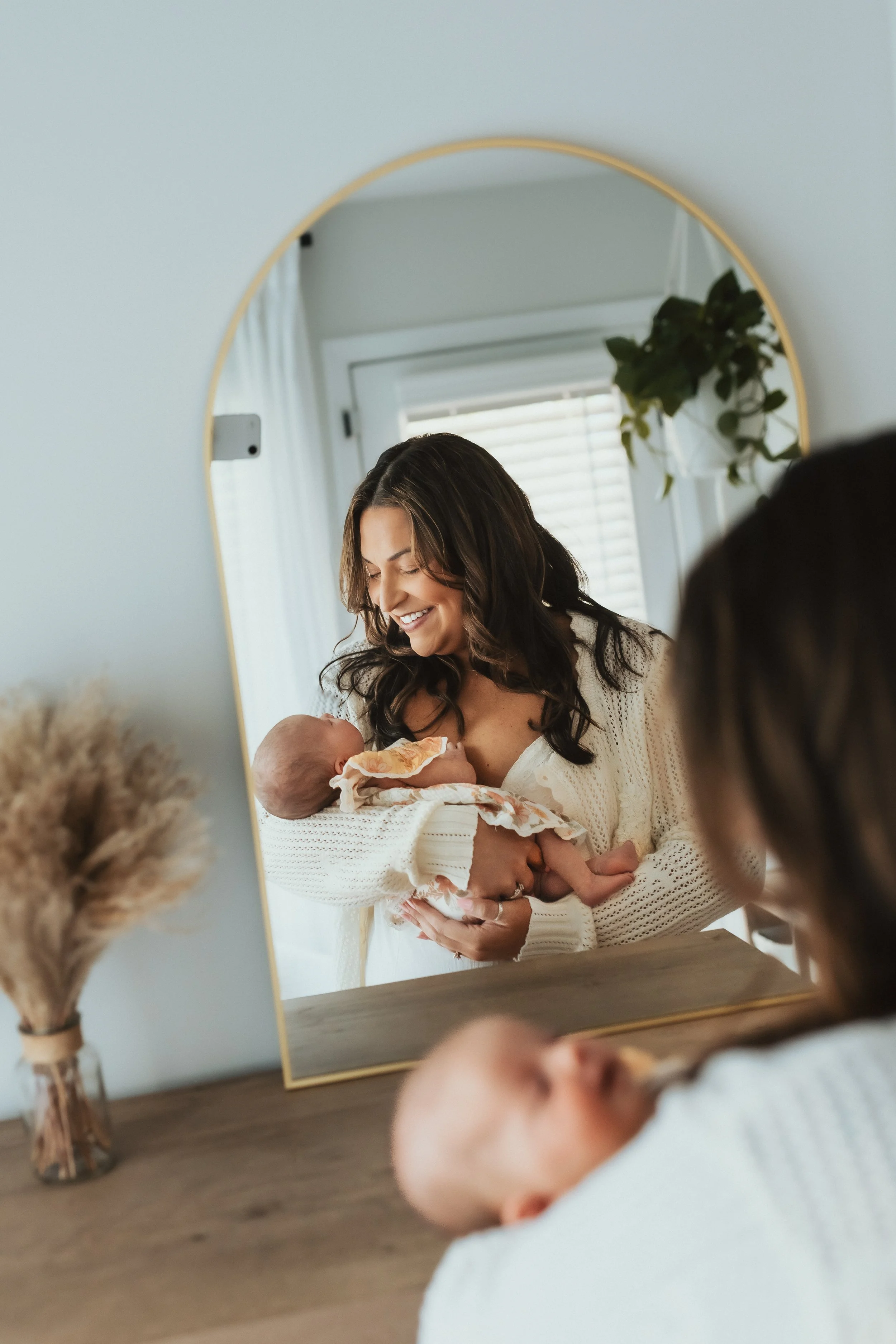A woman holding a baby in front of a mirror, smiling down at the baby. The mirror reflects a window with blinds and a small potted plant. The room has a warm, cozy feel, with soft natural light.