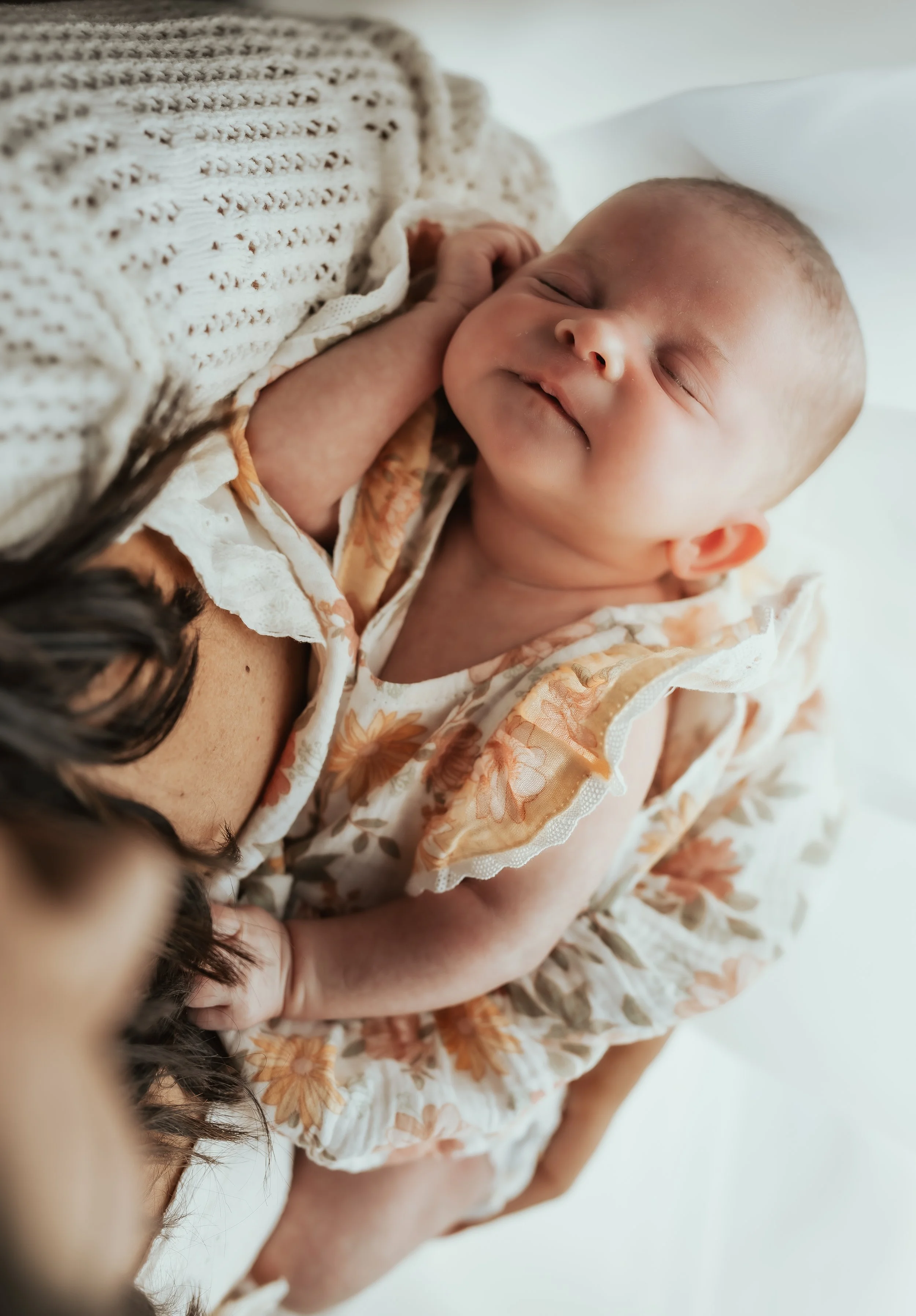 Close-up of a smiling baby in a floral outfit being held in a person's arms, with a knitted blanket nearby.