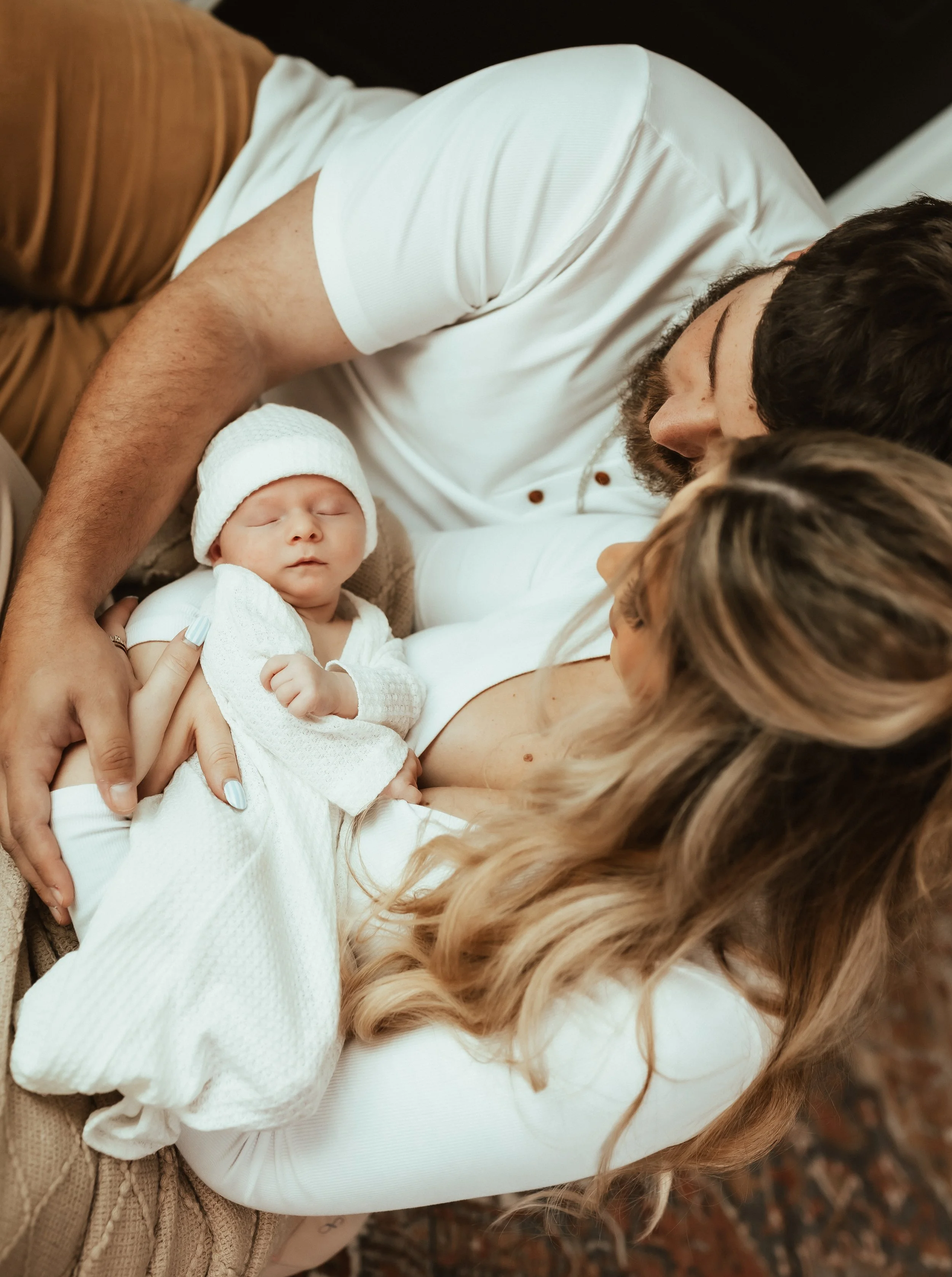 A woman laying on a bed with a man, holding a sleeping baby dressed in white with a hat.