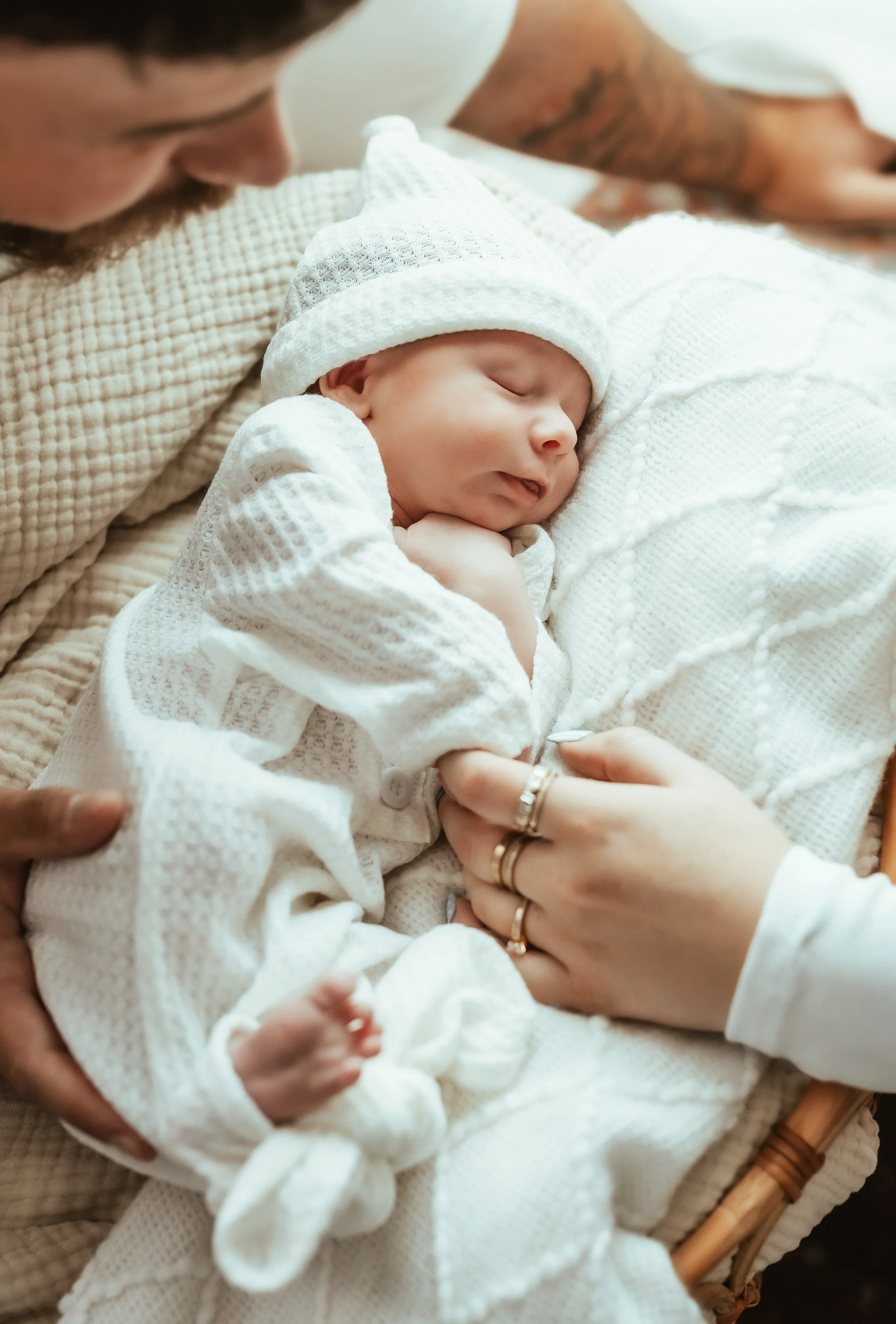 A sleeping baby dressed in a white knitted outfit and hat, resting on a soft white blanket, with a man in the background gently touching the baby.