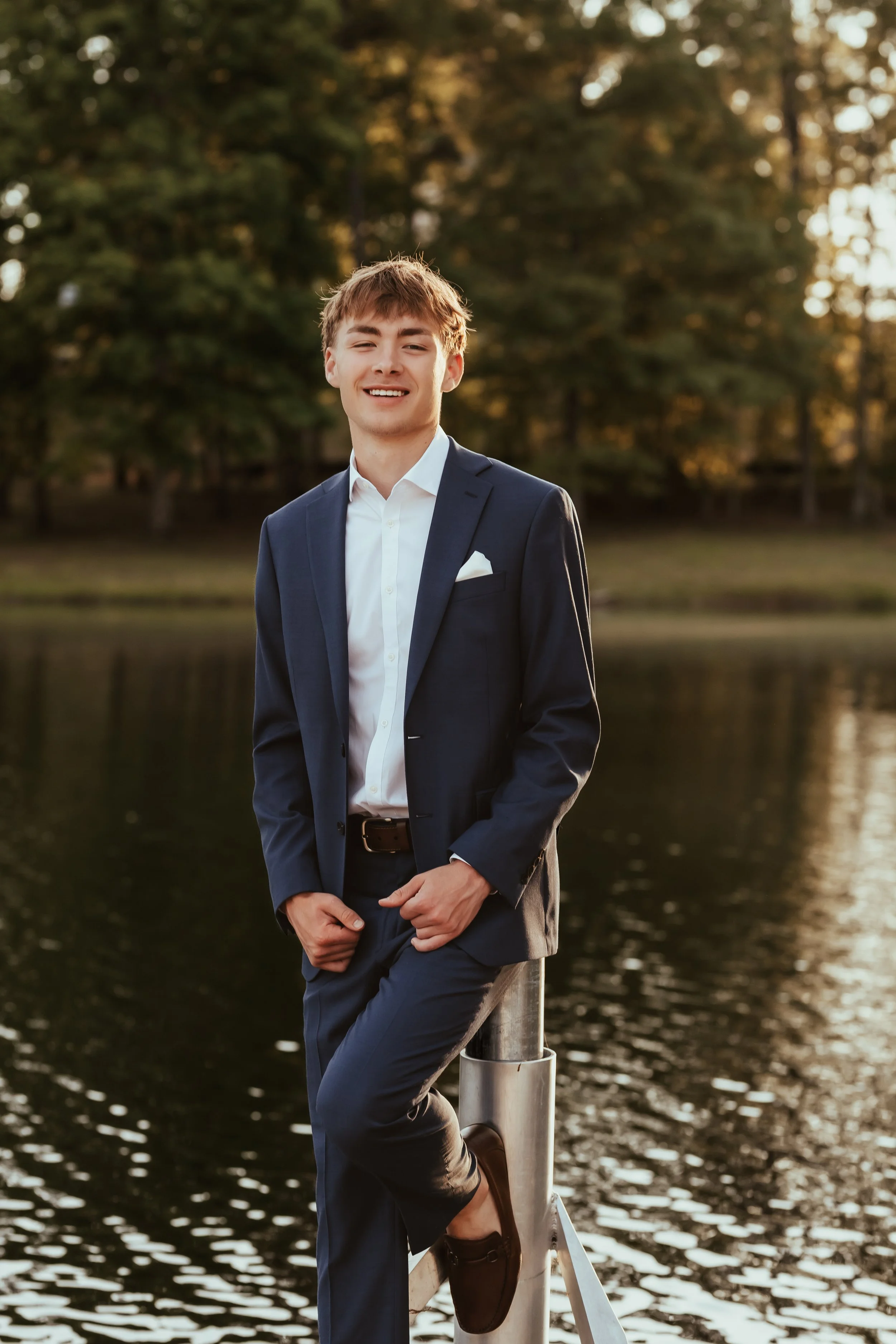 A young man in a navy suit and white shirt standing by a body of water during sunset, smiling, with trees and soft sunlight in the background.