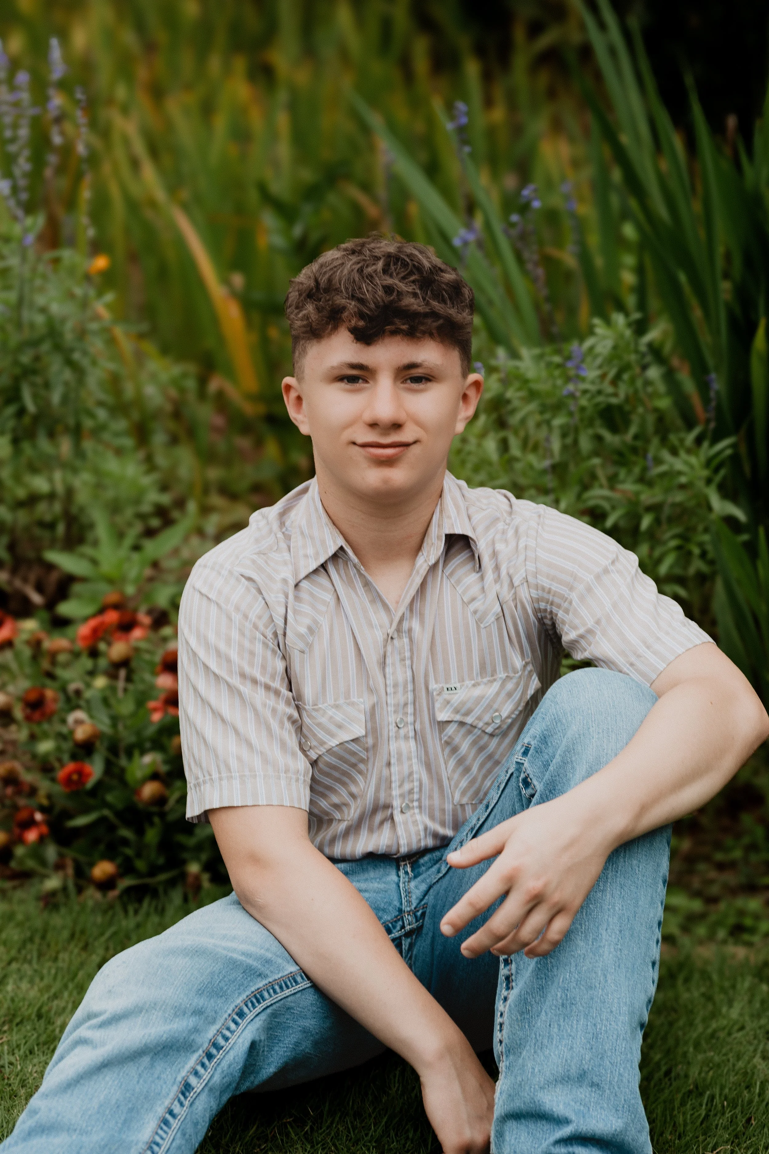 A young man with short, curly brown hair sitting outdoors in front of a lush green background of tall grass and purple flowers. He is wearing a beige and white striped short-sleeve button-up shirt and light blue jeans, and is smiling at the camera.